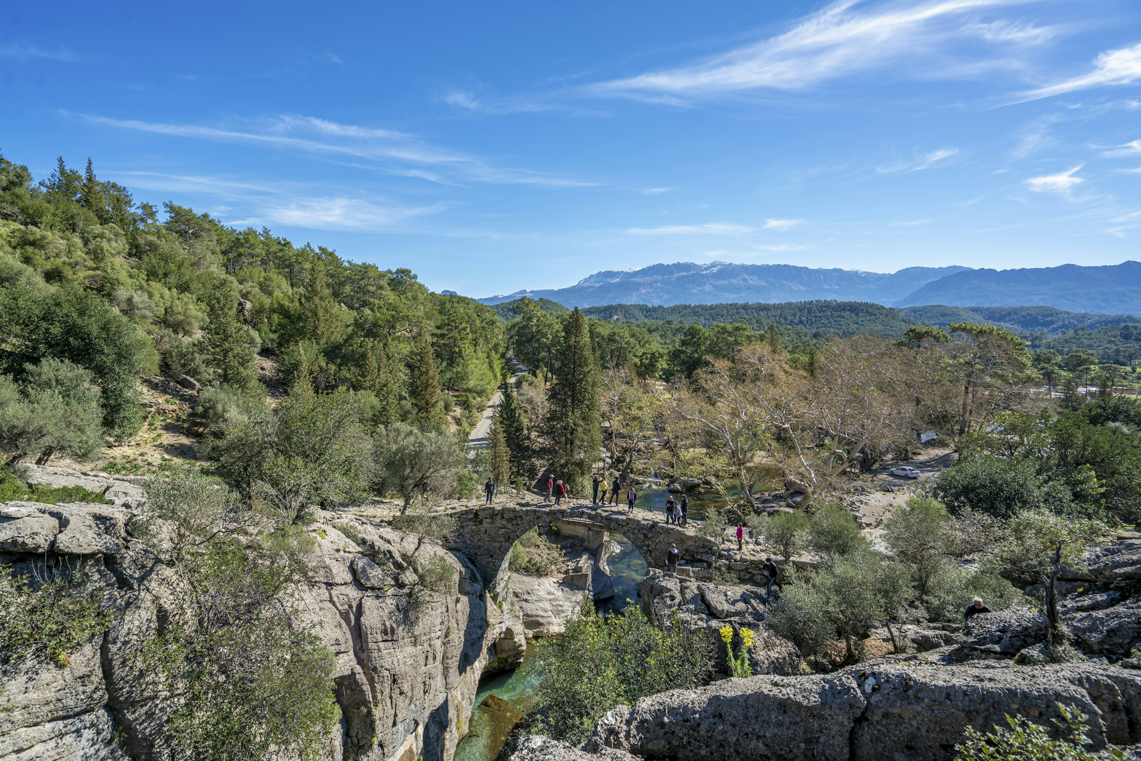 A group of hikers cross a small stone bridge across a river in Koprulu Kanyon Milli Parki. The bridge spanning the blue river is surrounded by forest in all directions.