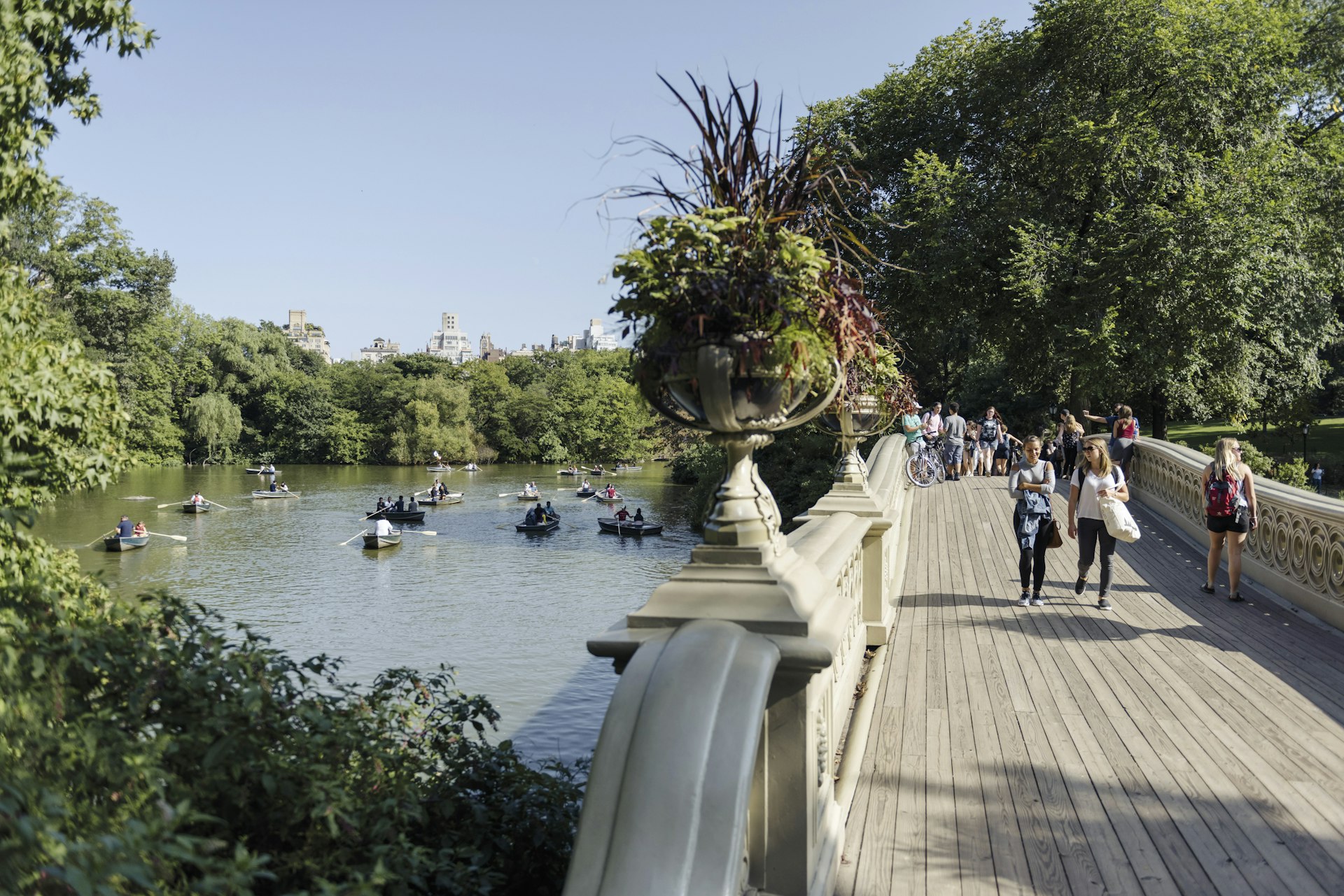 Row boats fill a lake in Central Park, as pedestrians cross a nearby bridge