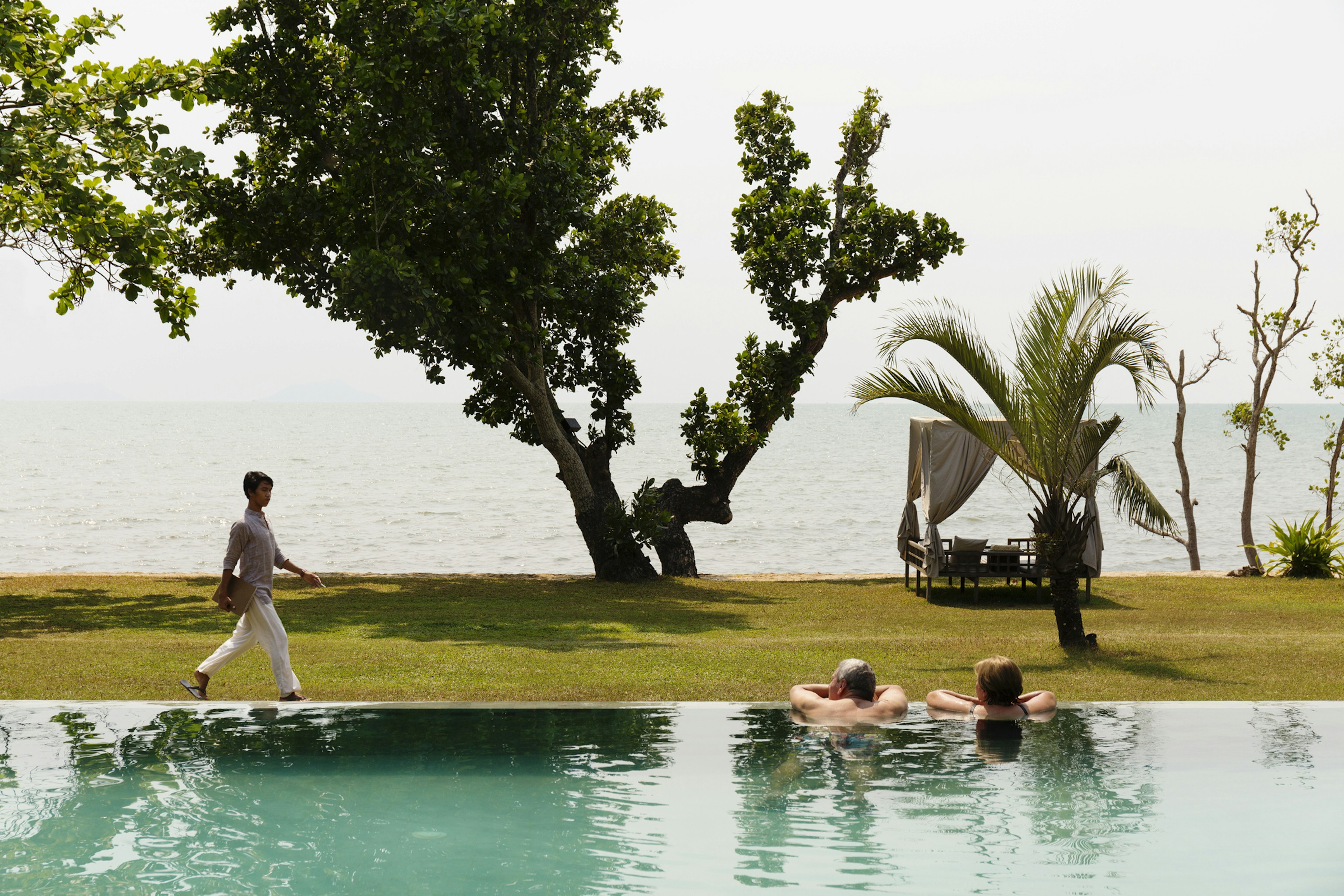 A couple lean on the edge of a swimming pool looking out towards the ocean. A woman is walking by