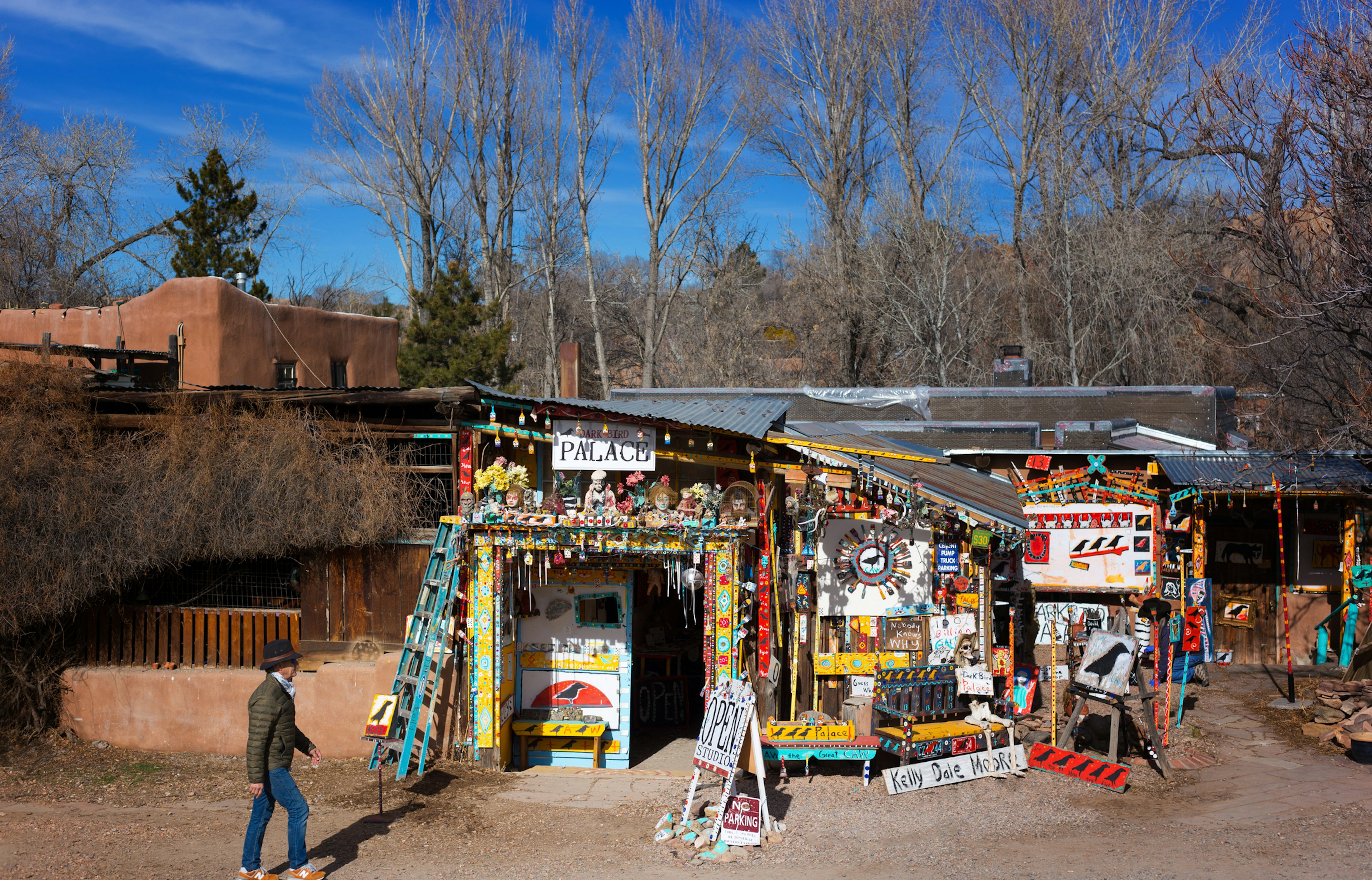 A man walking past an gallery filled with colorful pieces on Canyon Road, a famous street full of shops and art galleries in Santa Fe, New Mexico. Bare trees and adobe dwellings are visible in the background.