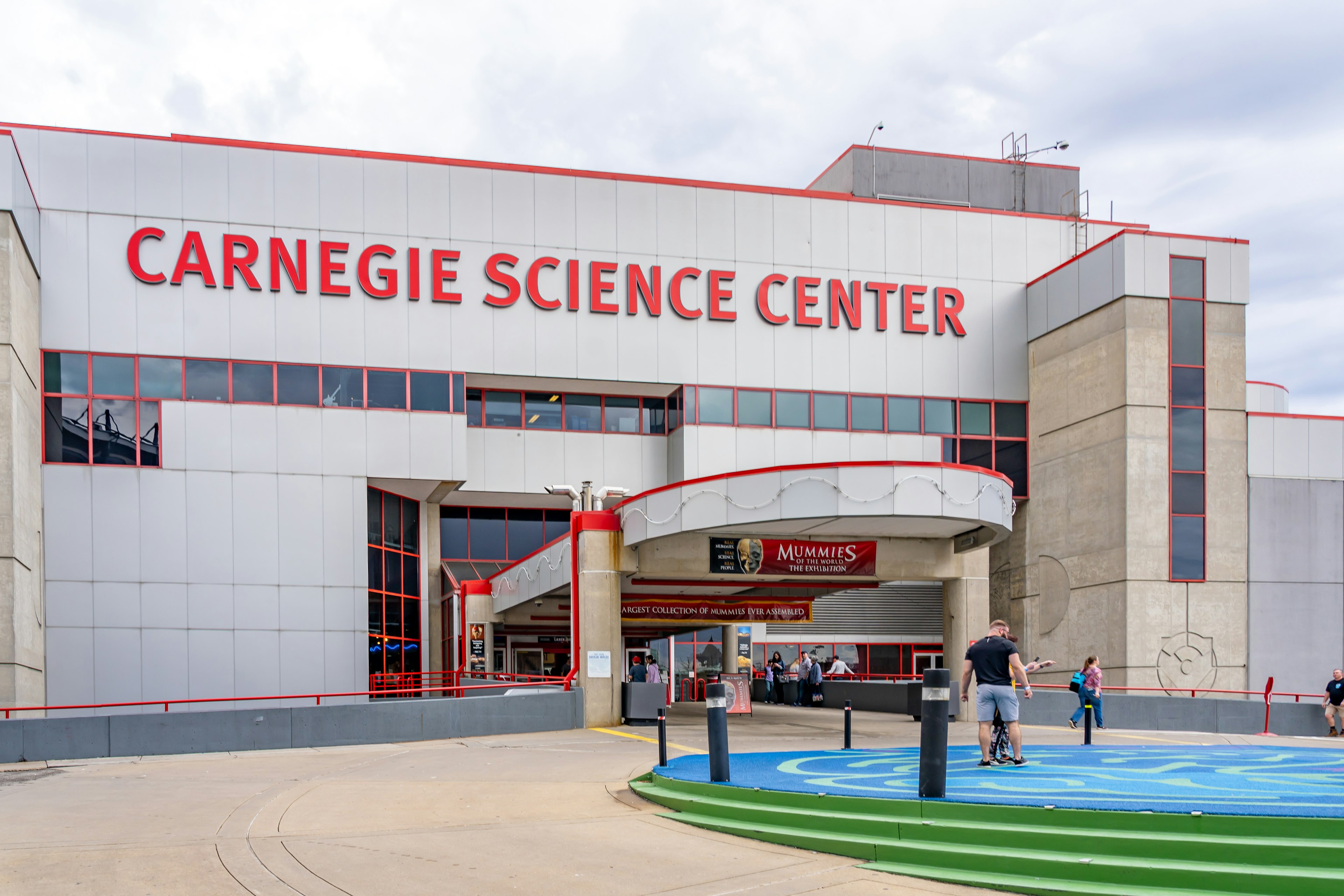 Entrance of Carnegie Science Center in Pittsburgh, Pennsylvania There are a few people standing outside of the red and gray building