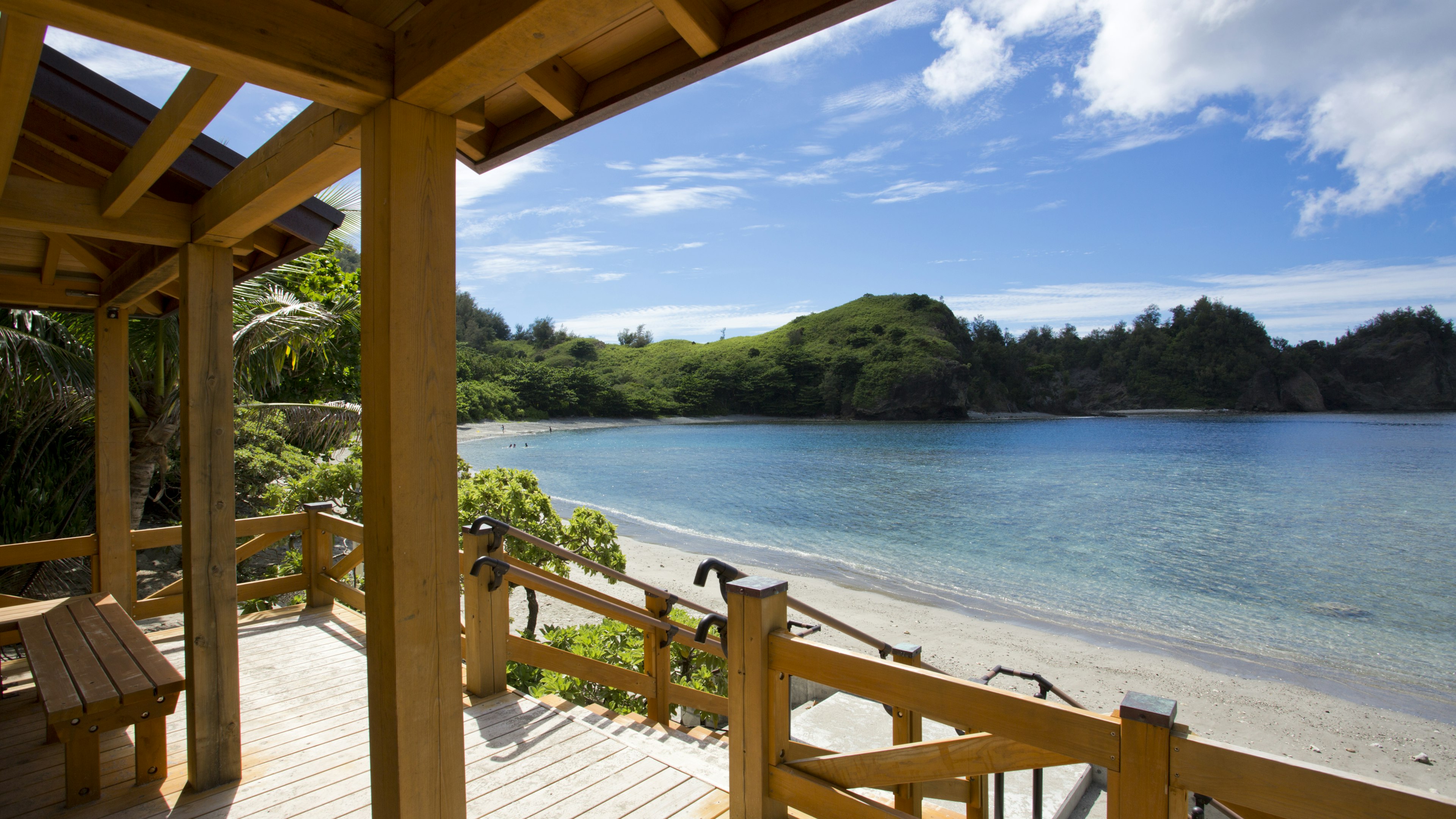 A view of Miyanohama beach on Chichi-jima island. The beach is a crescent-shaped stretch of golden sand with blue water, and is viewed in the picture from a wooden terrace of what is probably a beach bar.