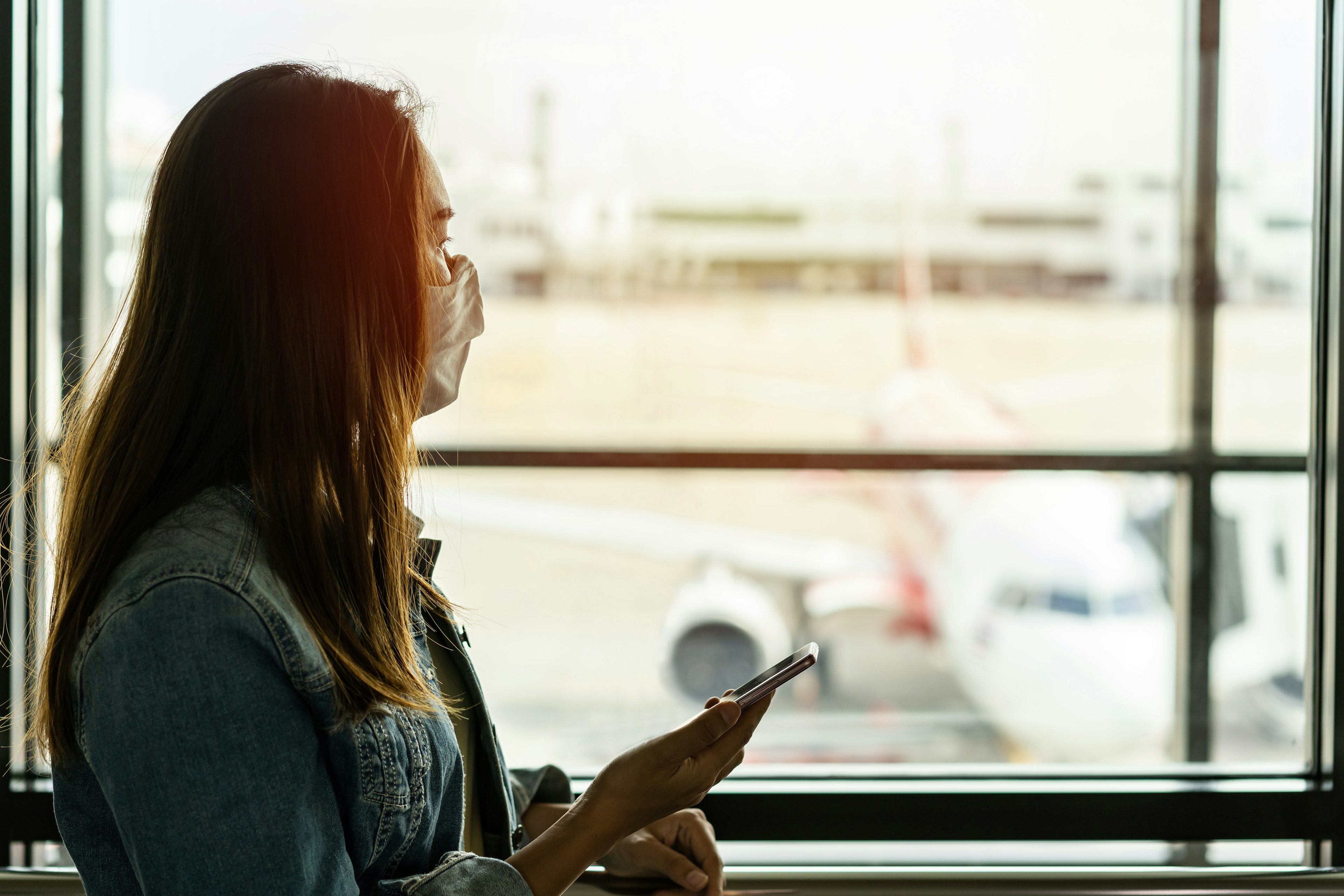 A woman wearing a mask stands near a window in the airport.