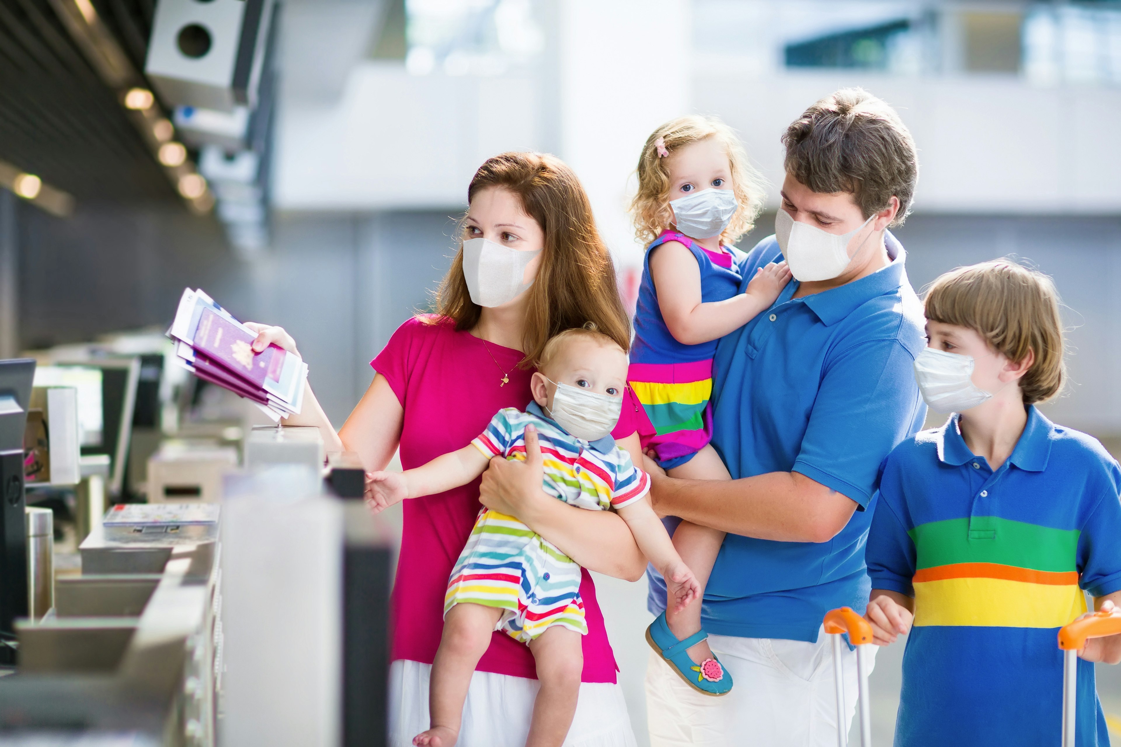 A white family of two adults and three young children, wearing brightly colored clothes, hold out their passports at an airport counter. They are all wearing face masks