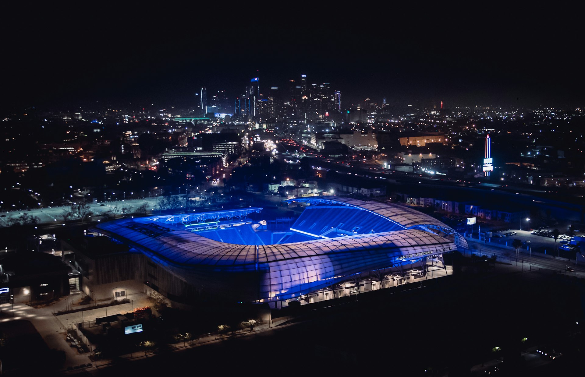 BMO Stadium illuminated with purple lights in an aerial view with the Los Angeles skyline in the distance