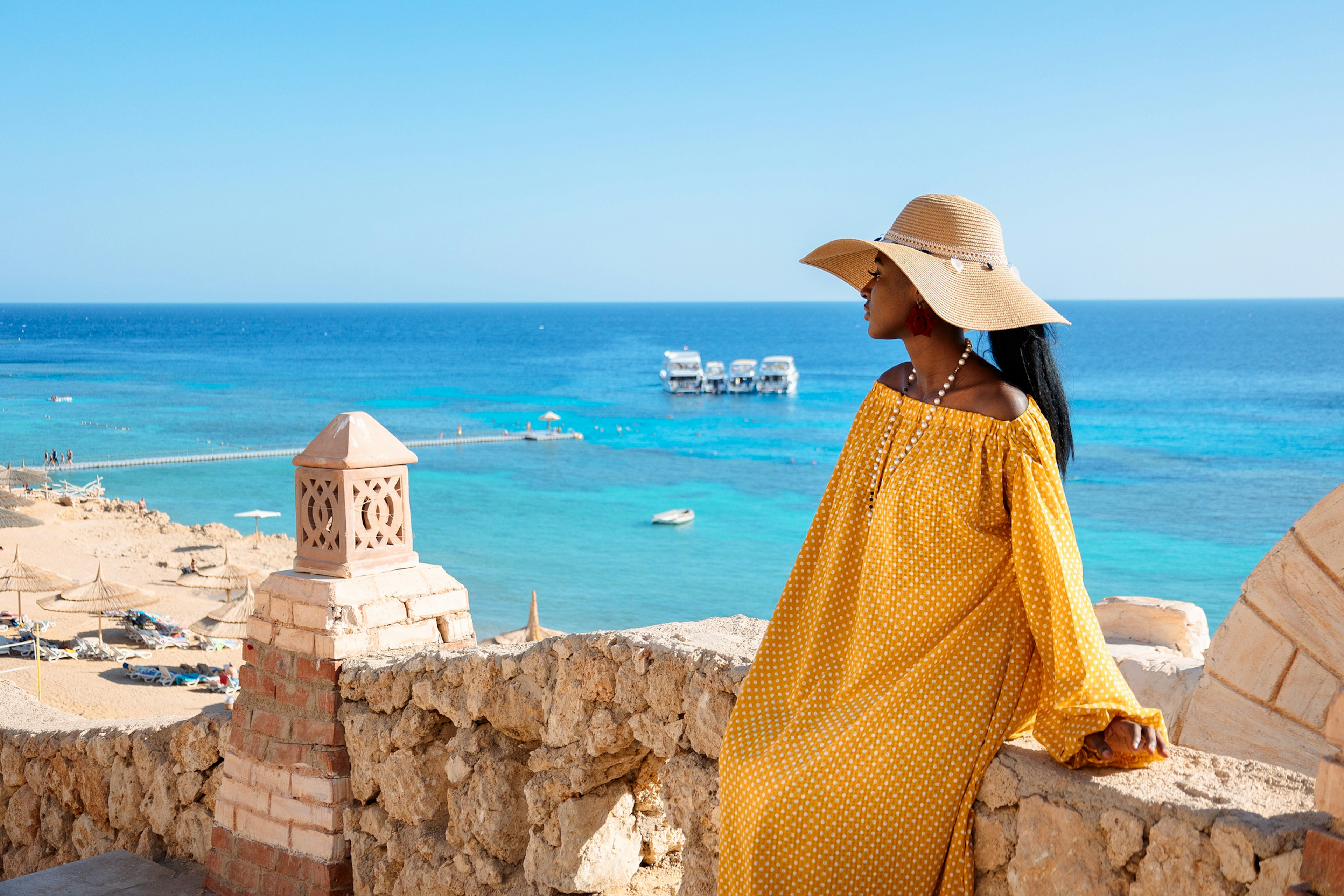 A woman wearing a yellow dress and a sunhat stands at an overlook above a sandy beach and turquoise sea in Sharm El Sheikh, Egypt