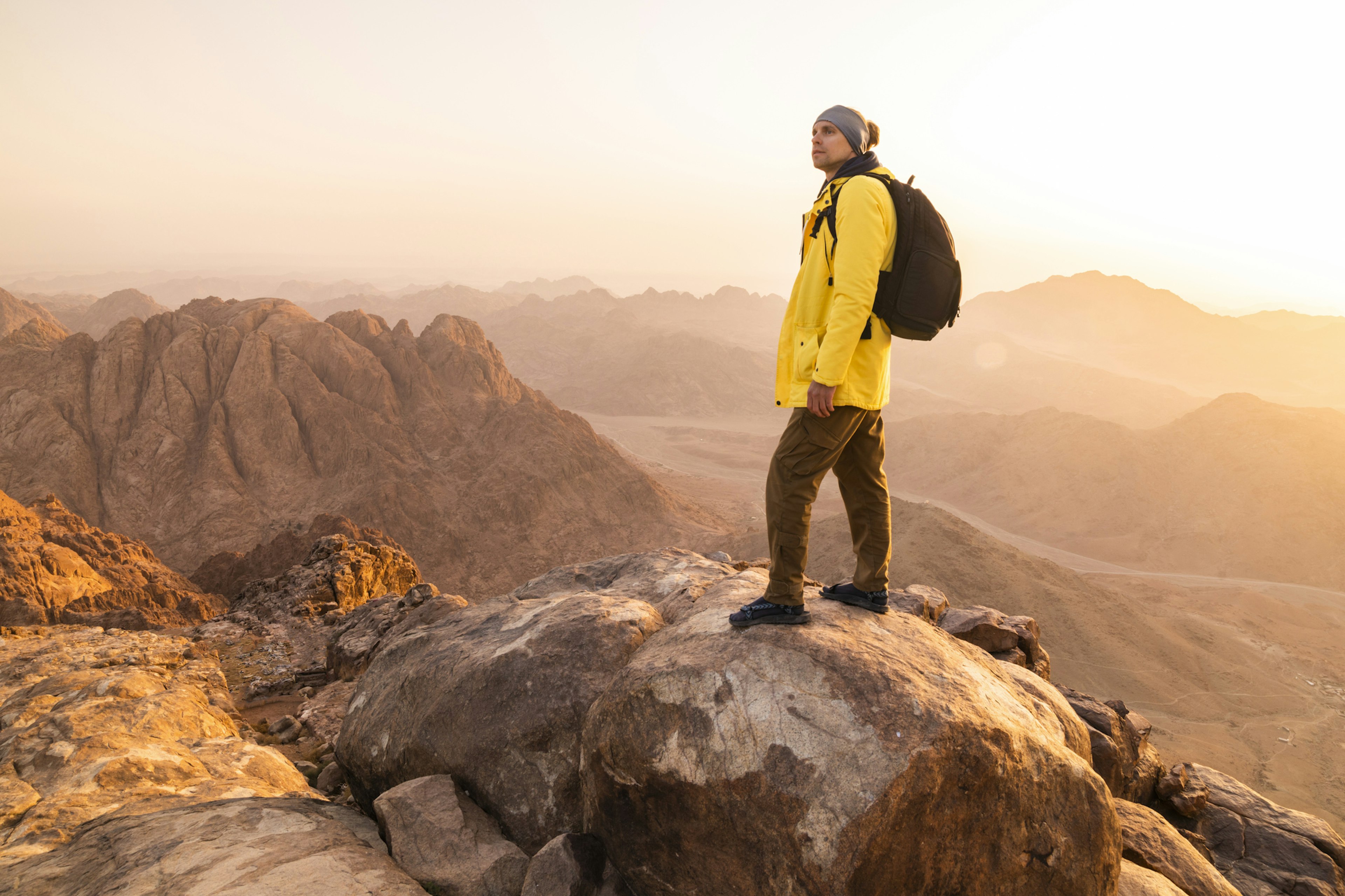 A hiker in a bright yellow coat with a black rucksack stands at the top of a mountain in Sinai, Egypt