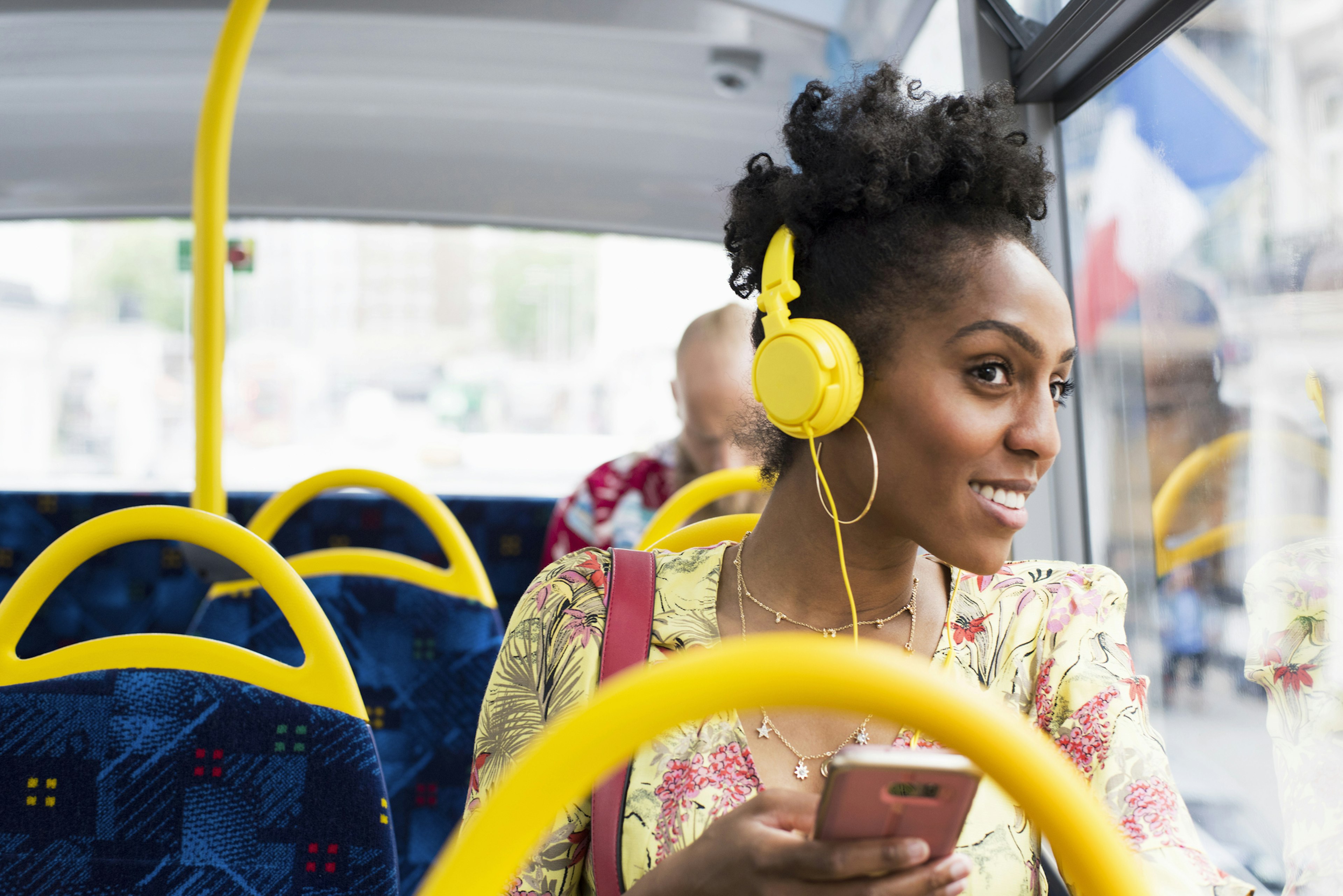 A black woman wearing headphones is looking out of the window of a bus and smiling