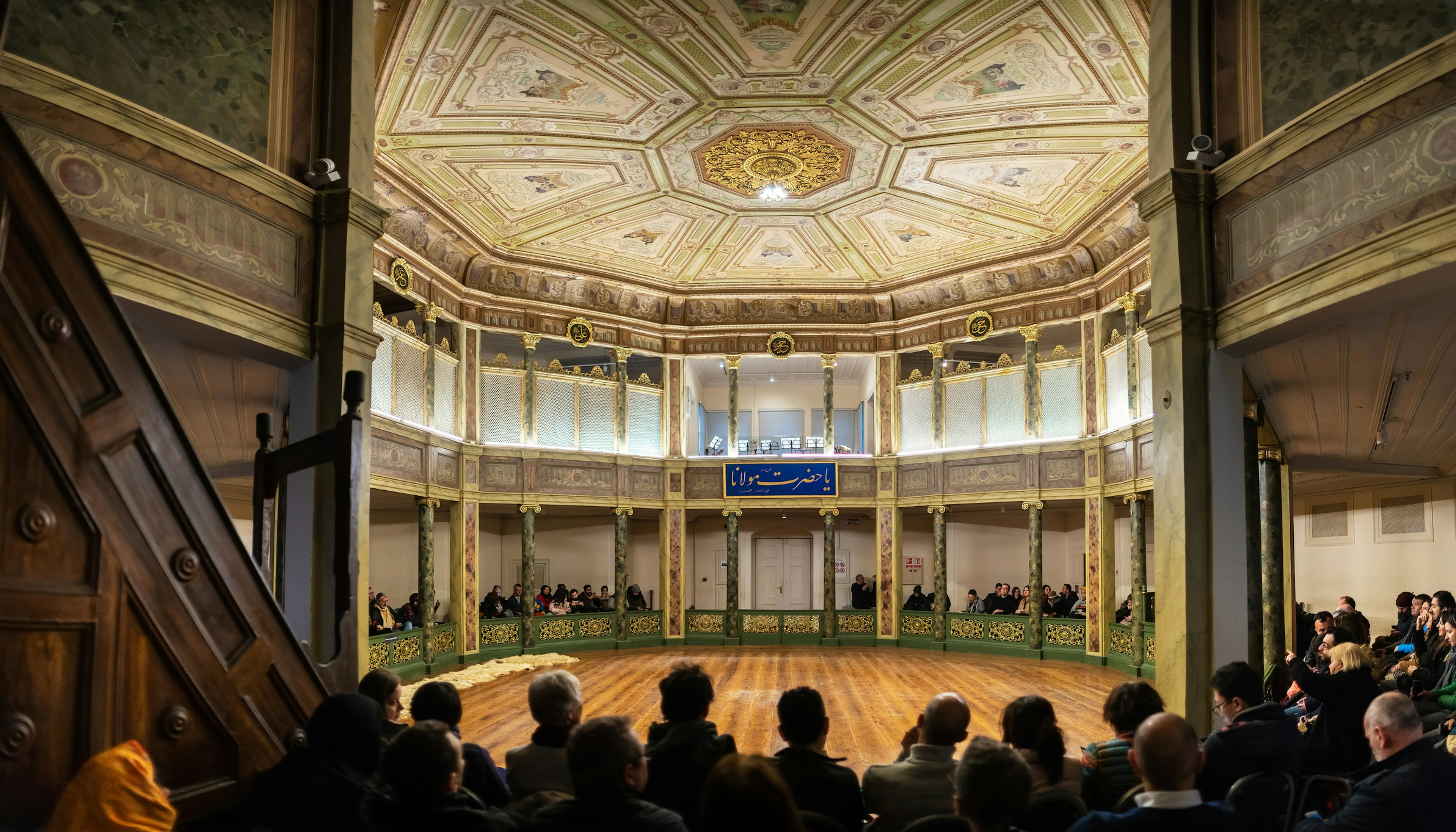 Tourists seated facing an empty round floor at the Galata Mevlevi House Museum