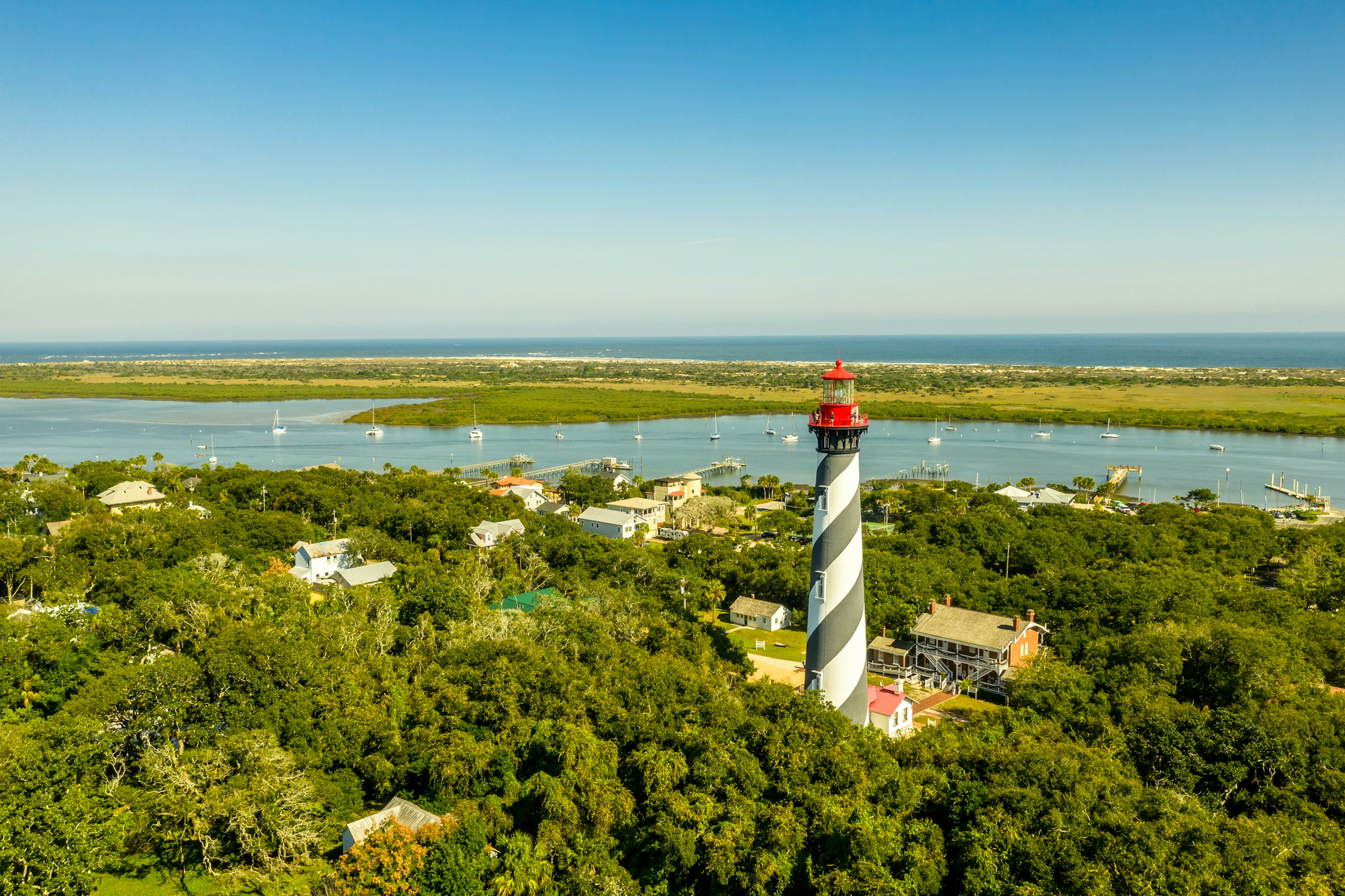 Aerial view of a black-and-white striped lighthouse. A peninsula in the distance covered in greenery gives way to a long stretch of golden sand