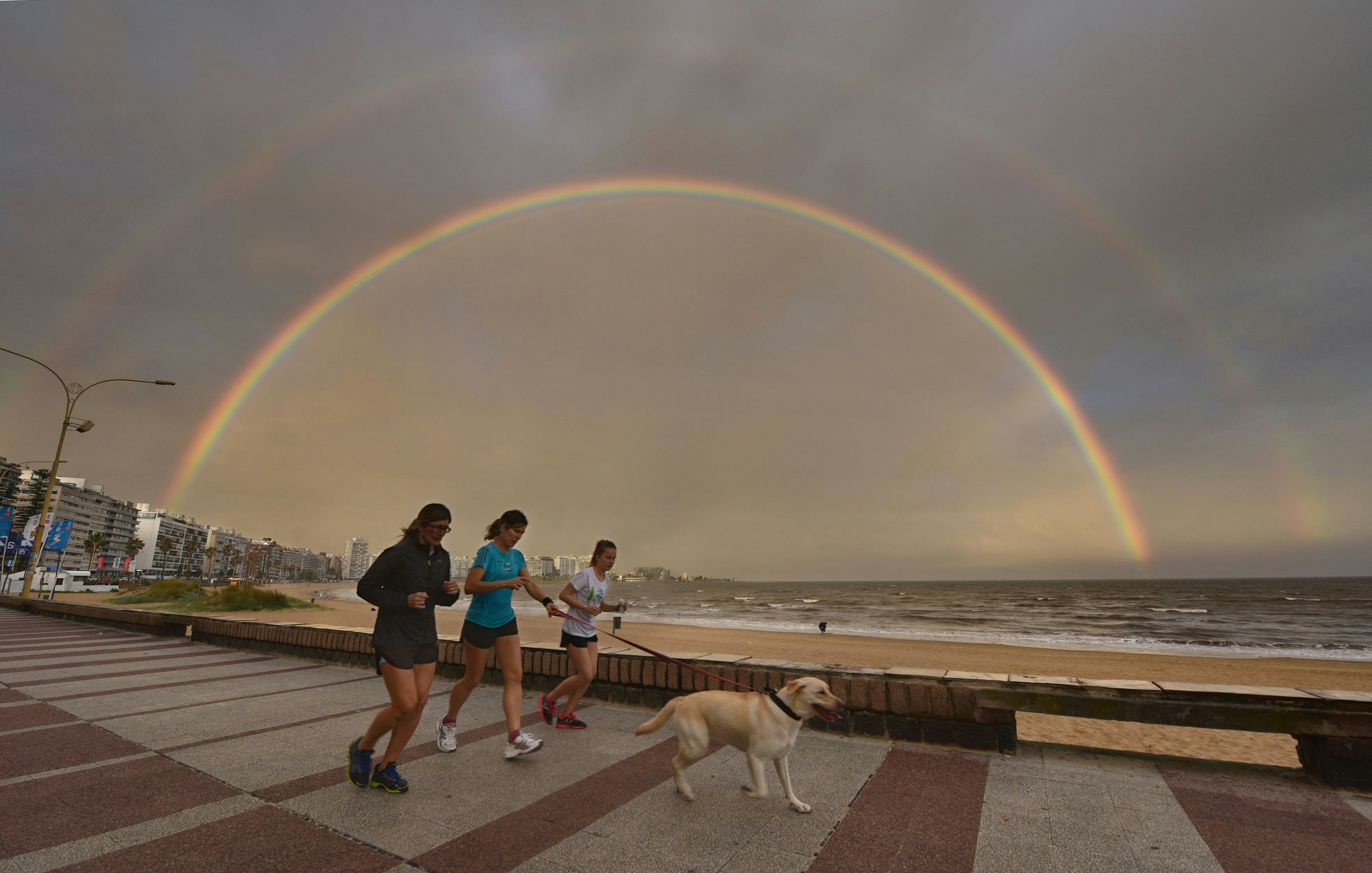 People jog with a dog along Pocitos' Rambla as a double rainbow appears during sunset over Montevideo