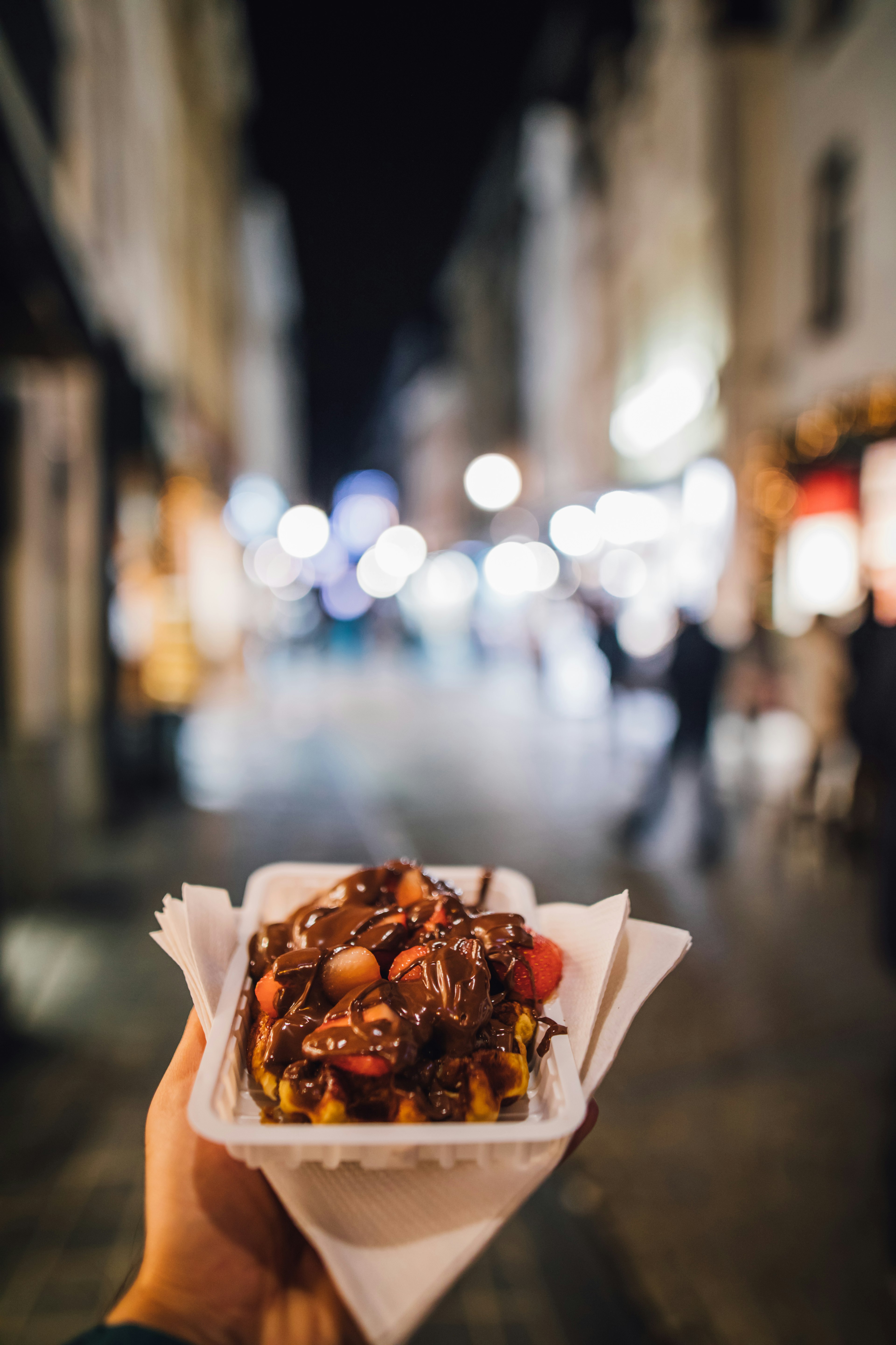 A person holds out a disposable plate containing a large Belgian waffle. The large sugary snack is smothered in chocolate sauce.