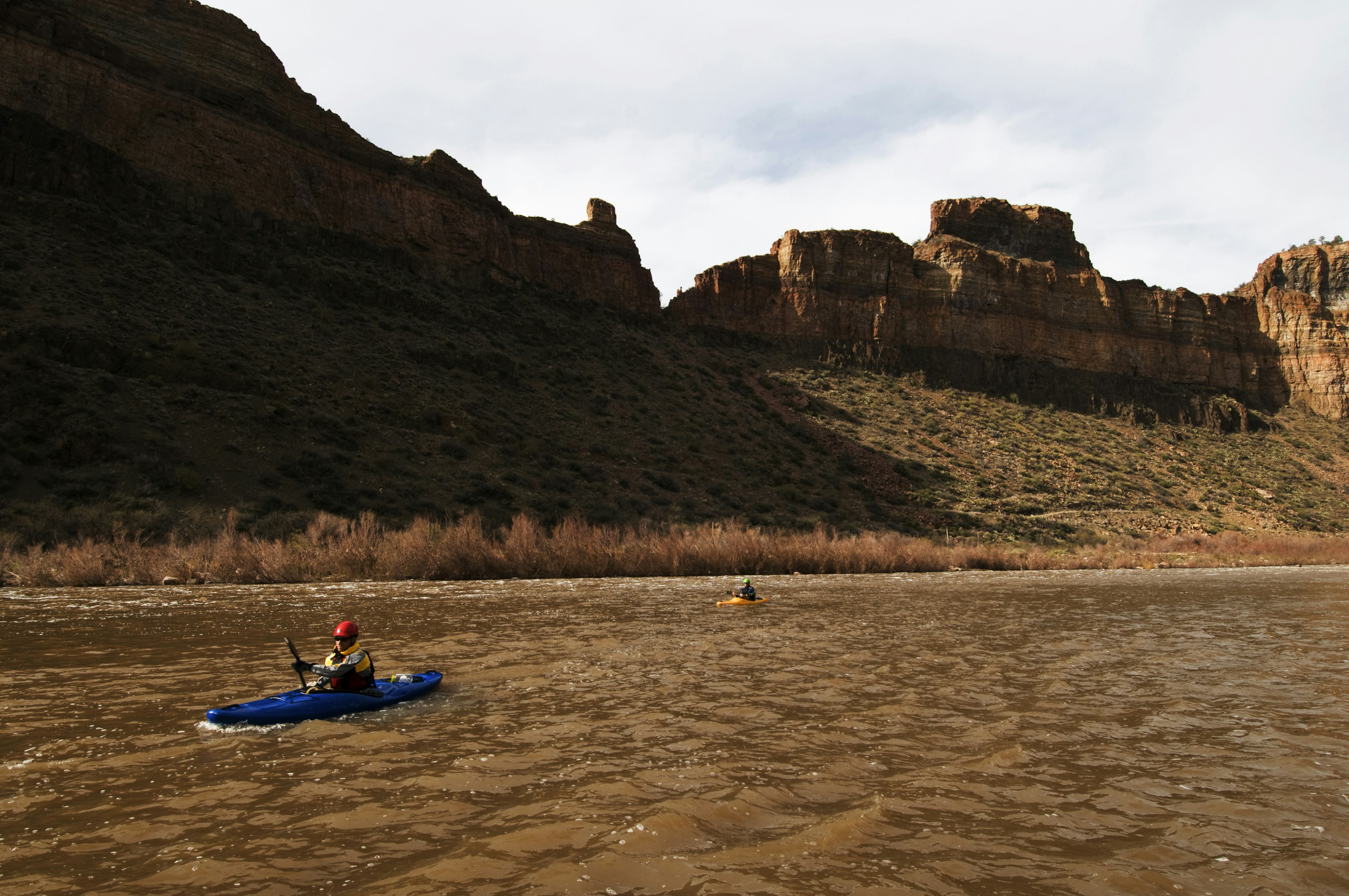 Kayakers paddle their boats on the Salt River against rocky mountains in shadow