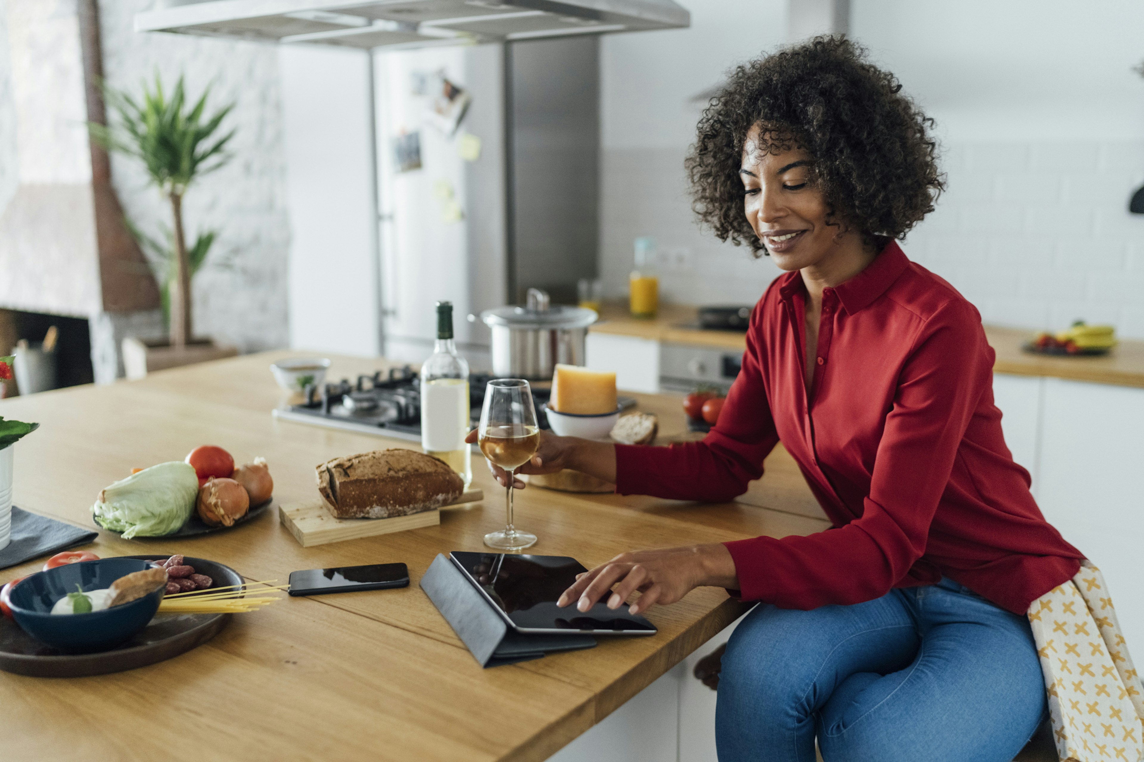 Woman sitting in kitchen with a glass of white wine, using digital tablet