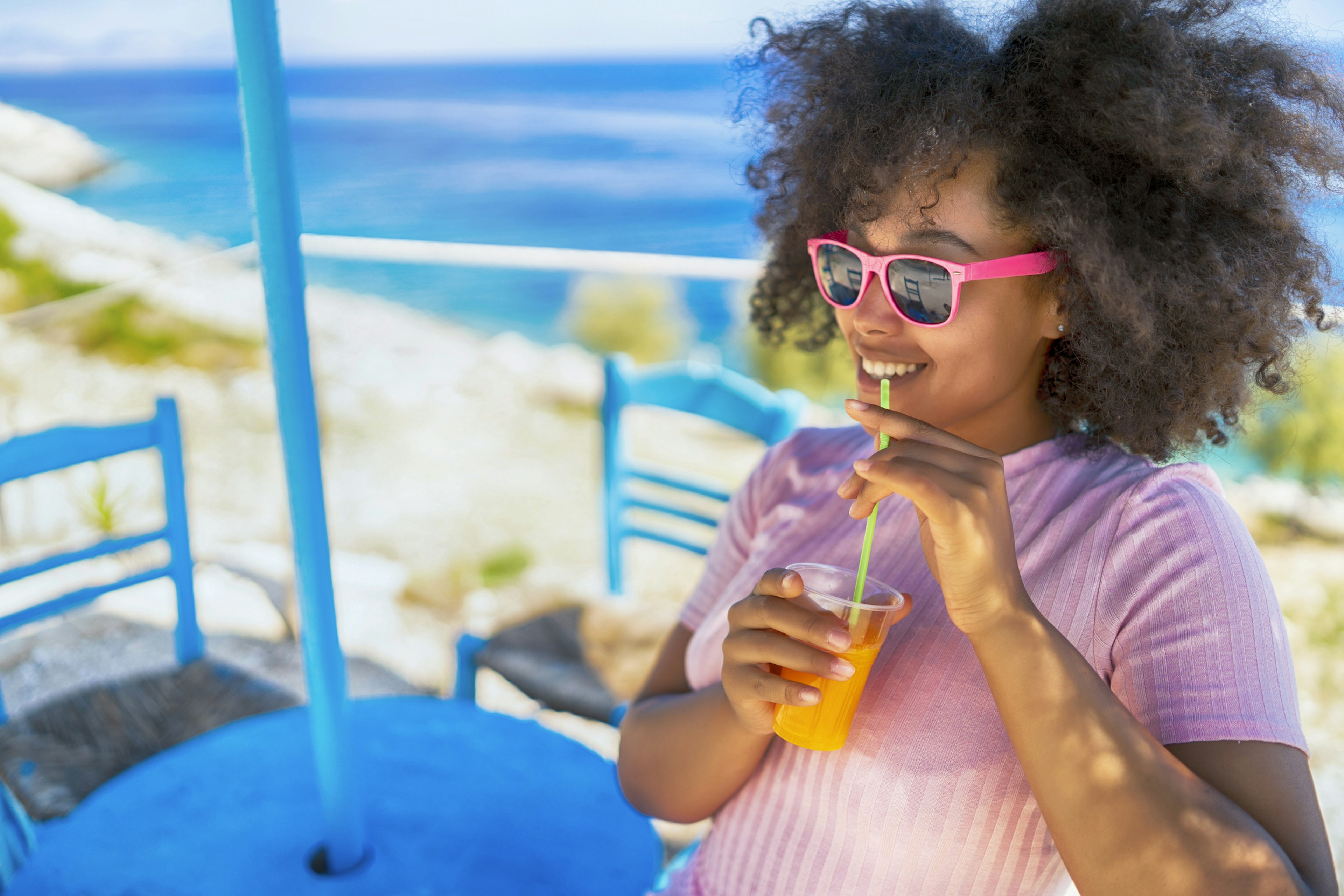 A black woman is sitting outside at a table in the sunshine, smiling and enjoying a drink