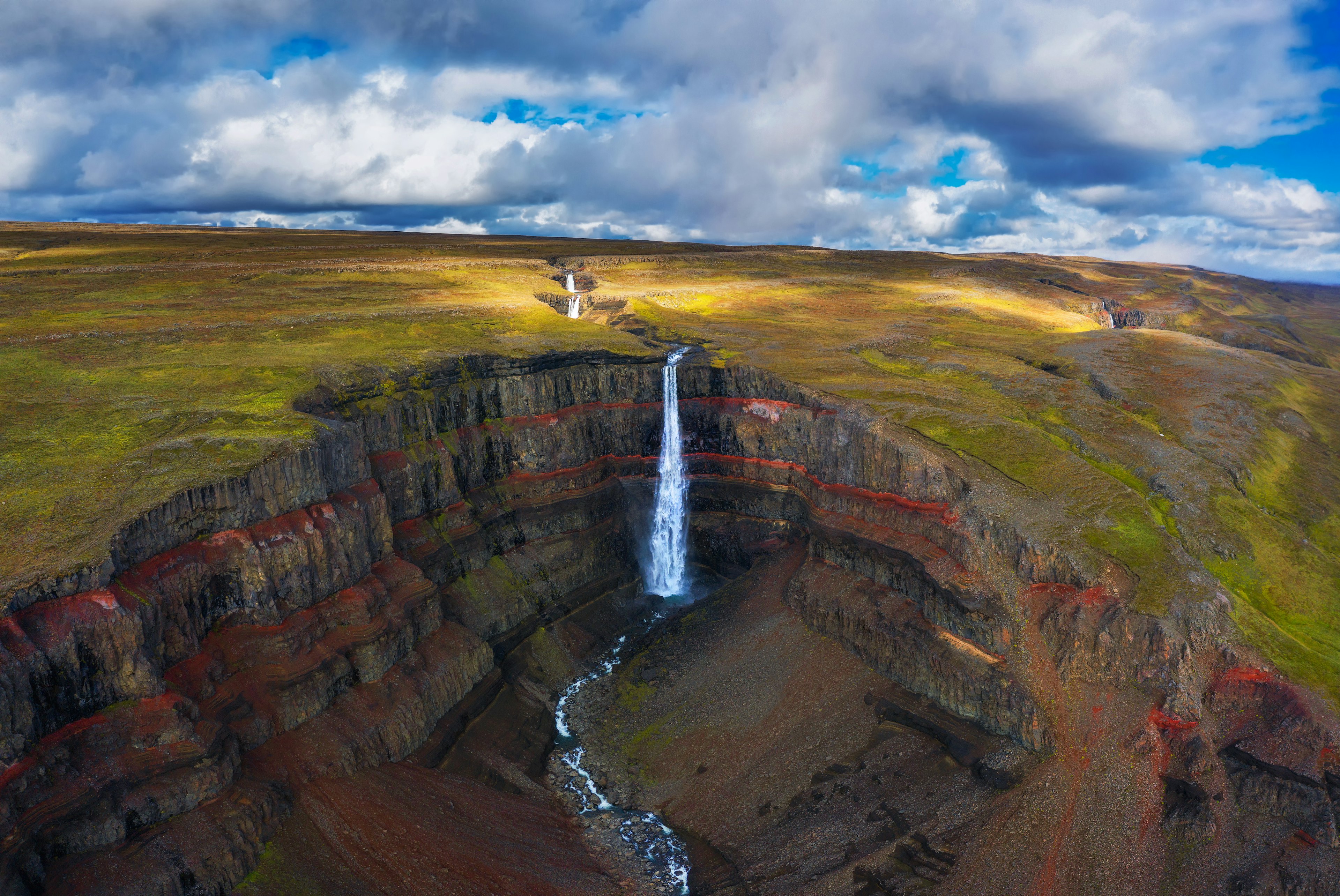 Aerial view of a high waterfall falling down into a crater with red layers of clay