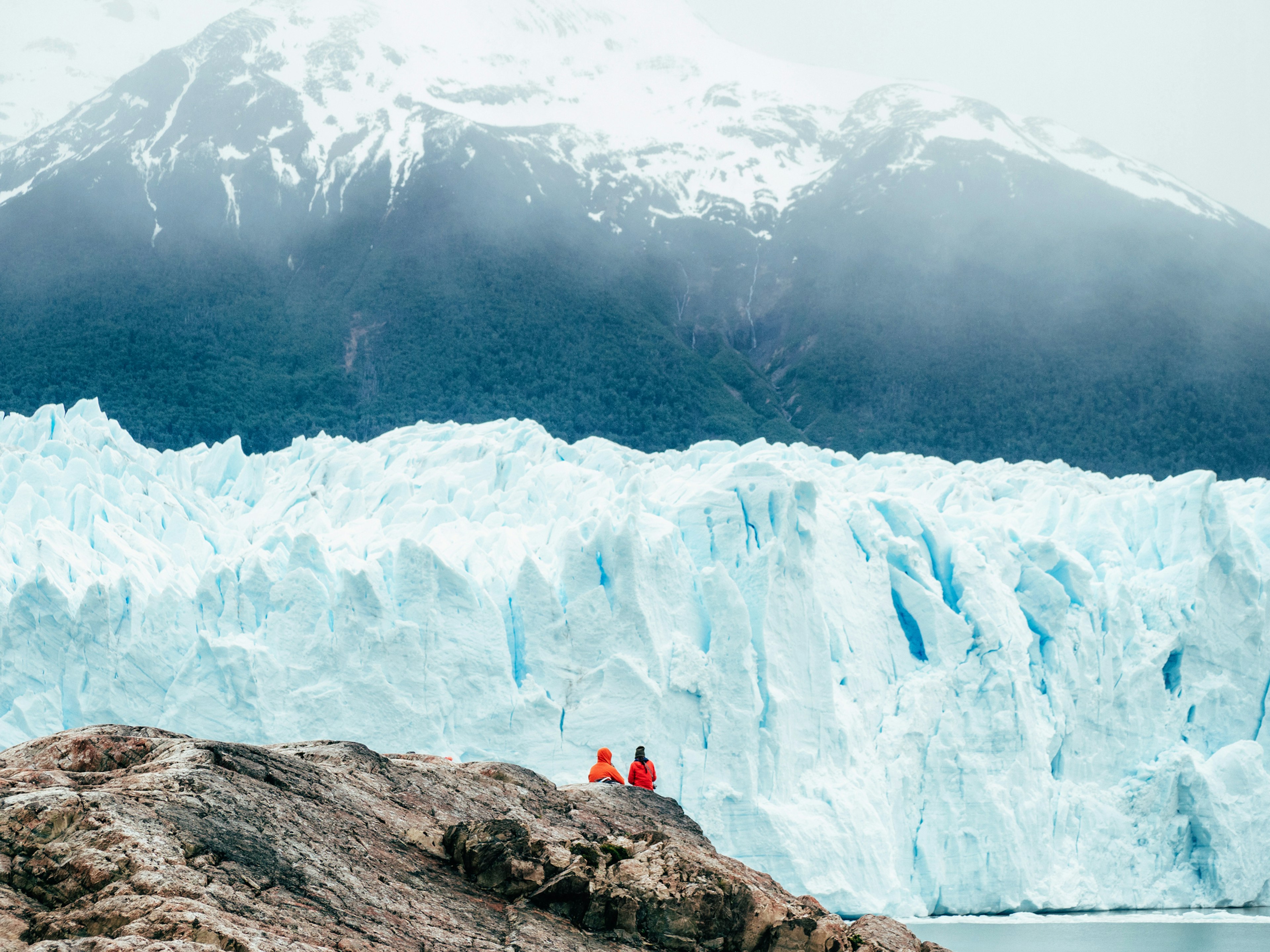 Two people sit at a viewpoint admiring a majestic blue-white wall of ice rising in front of them