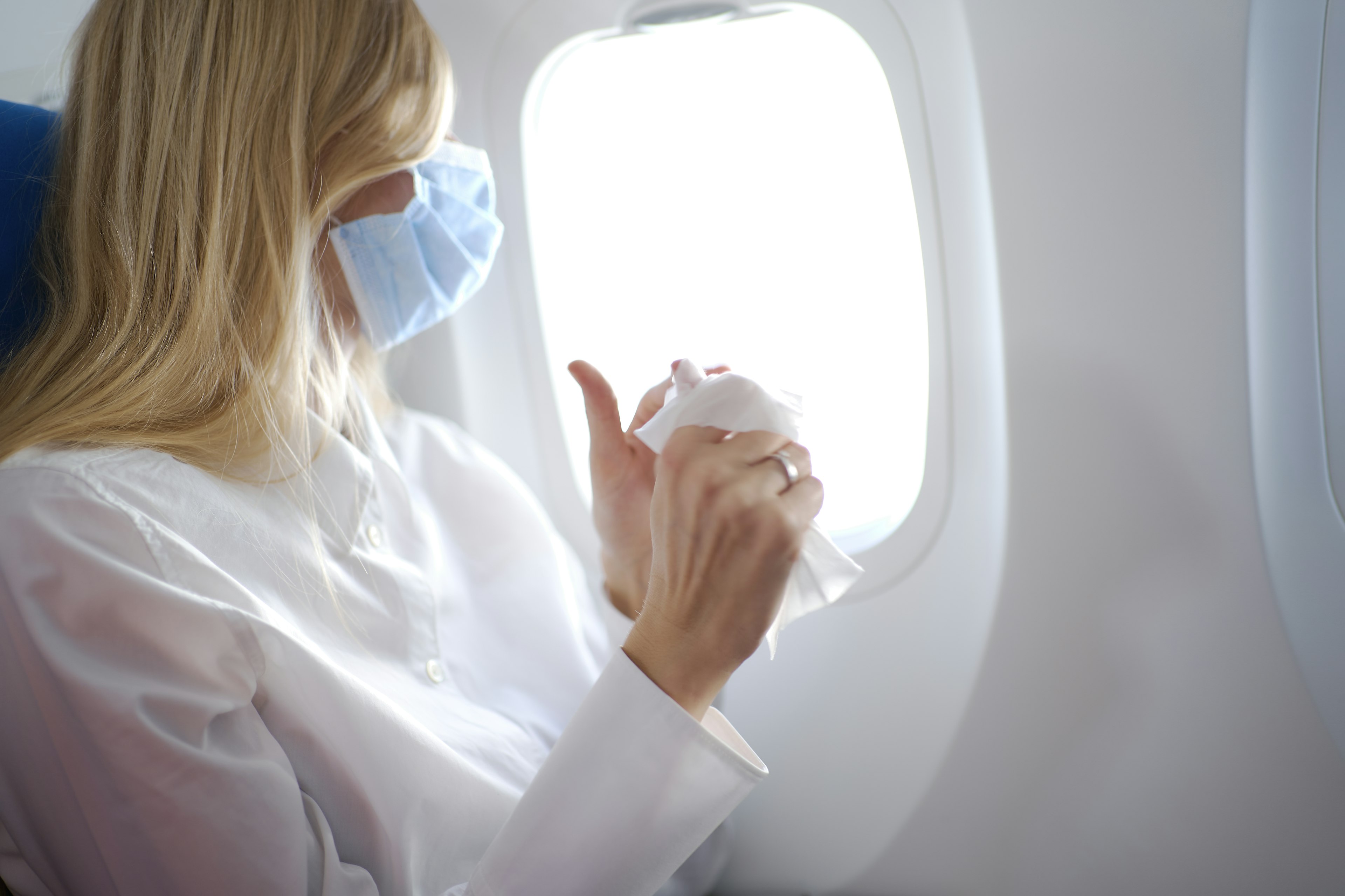 A woman with long blond hair wearing mask and white shirt inside airplane and cleaning her hands using wet wipes