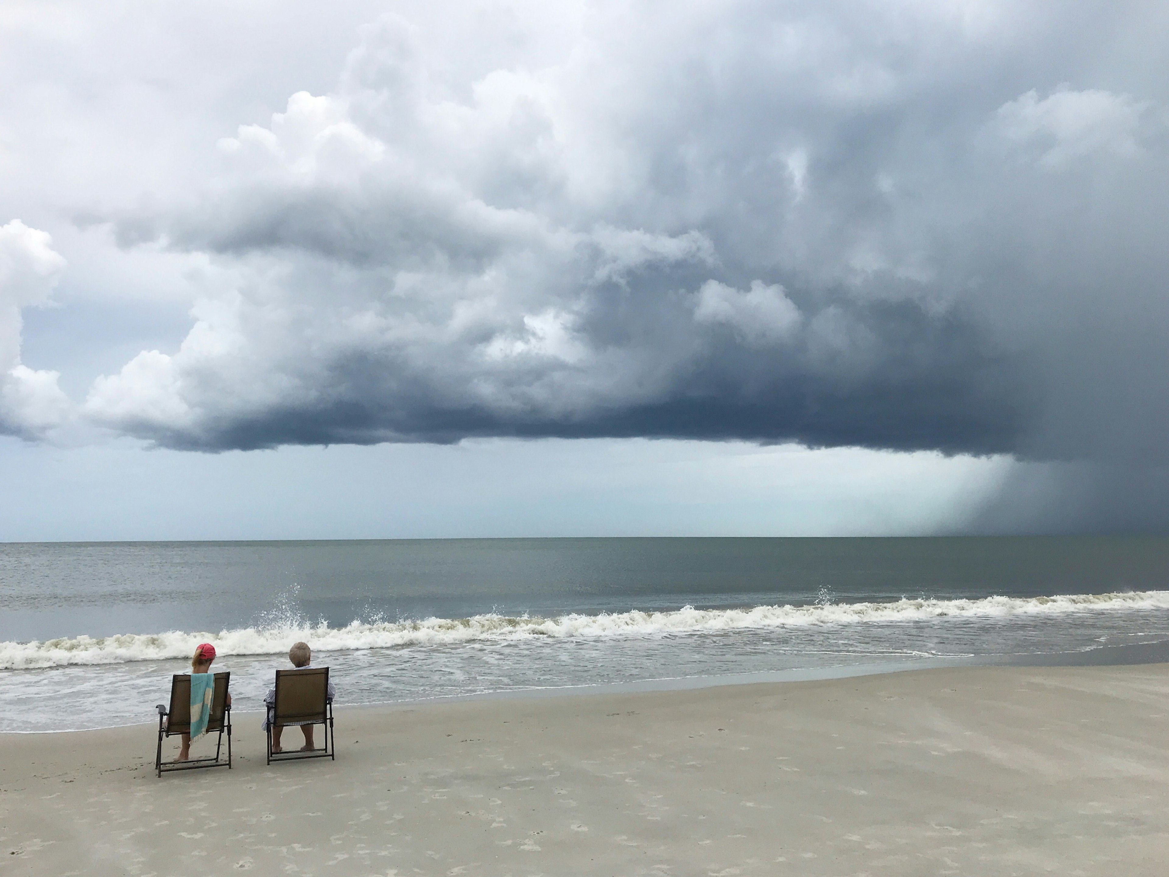 Tiny figures sit on beach chairs at the seaside beneath dramatic dark gray clouds swirling above as a storm front moves in