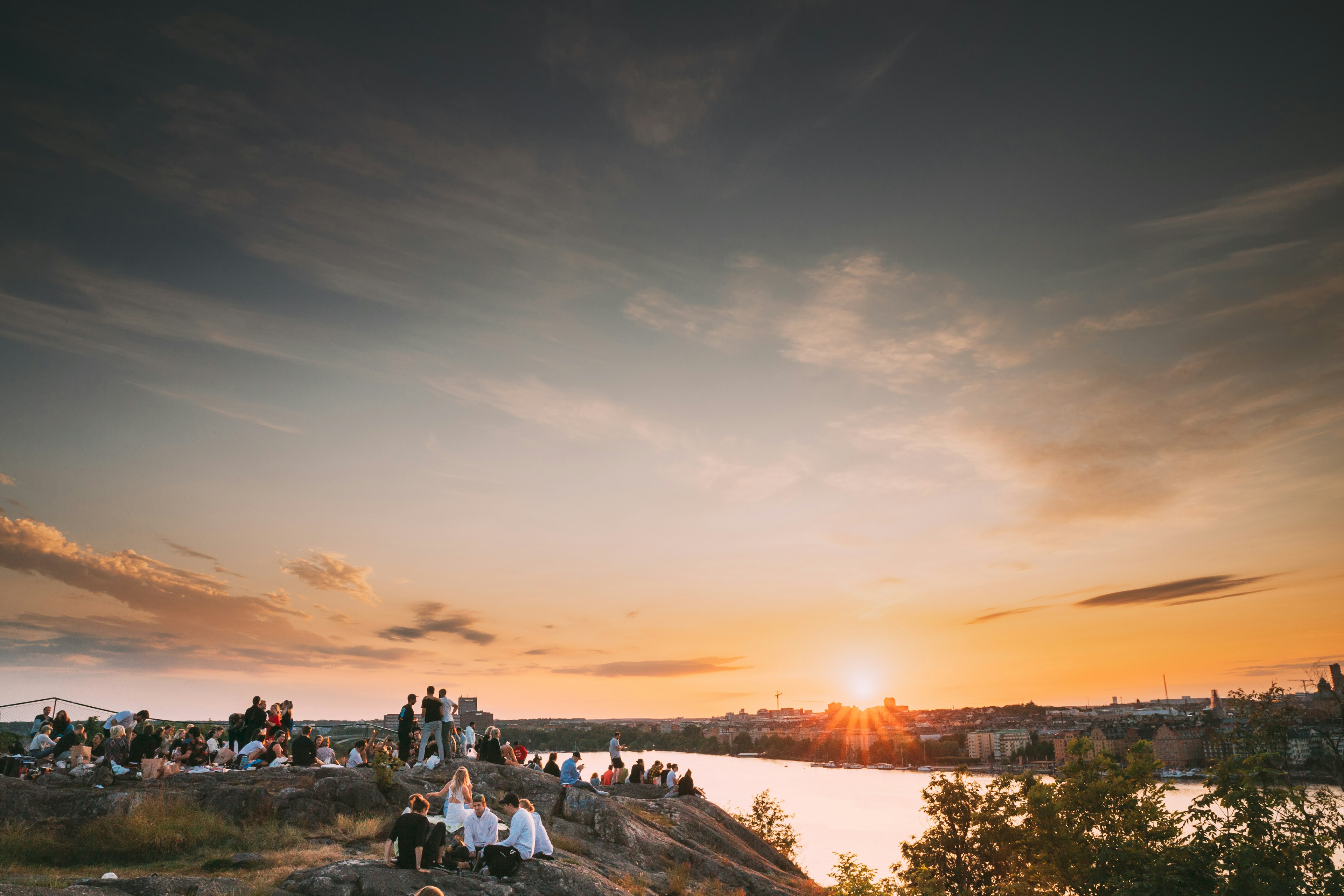 Groups of people sat on a cliff top looking out over a city at sunset