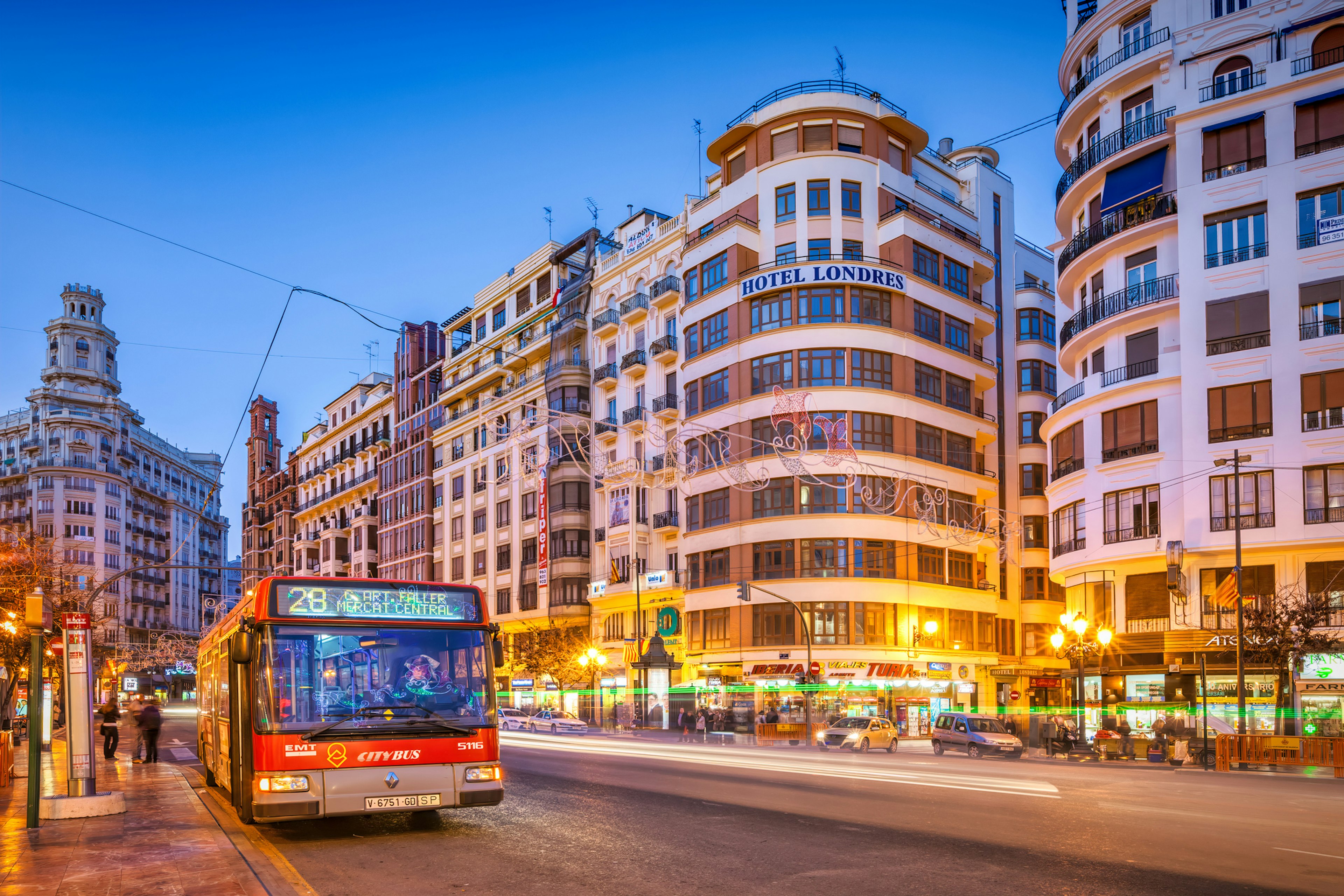 A red bus in downtown Valencia at dusk heads past a series of high-rise buildings which are bathed in illuminated light