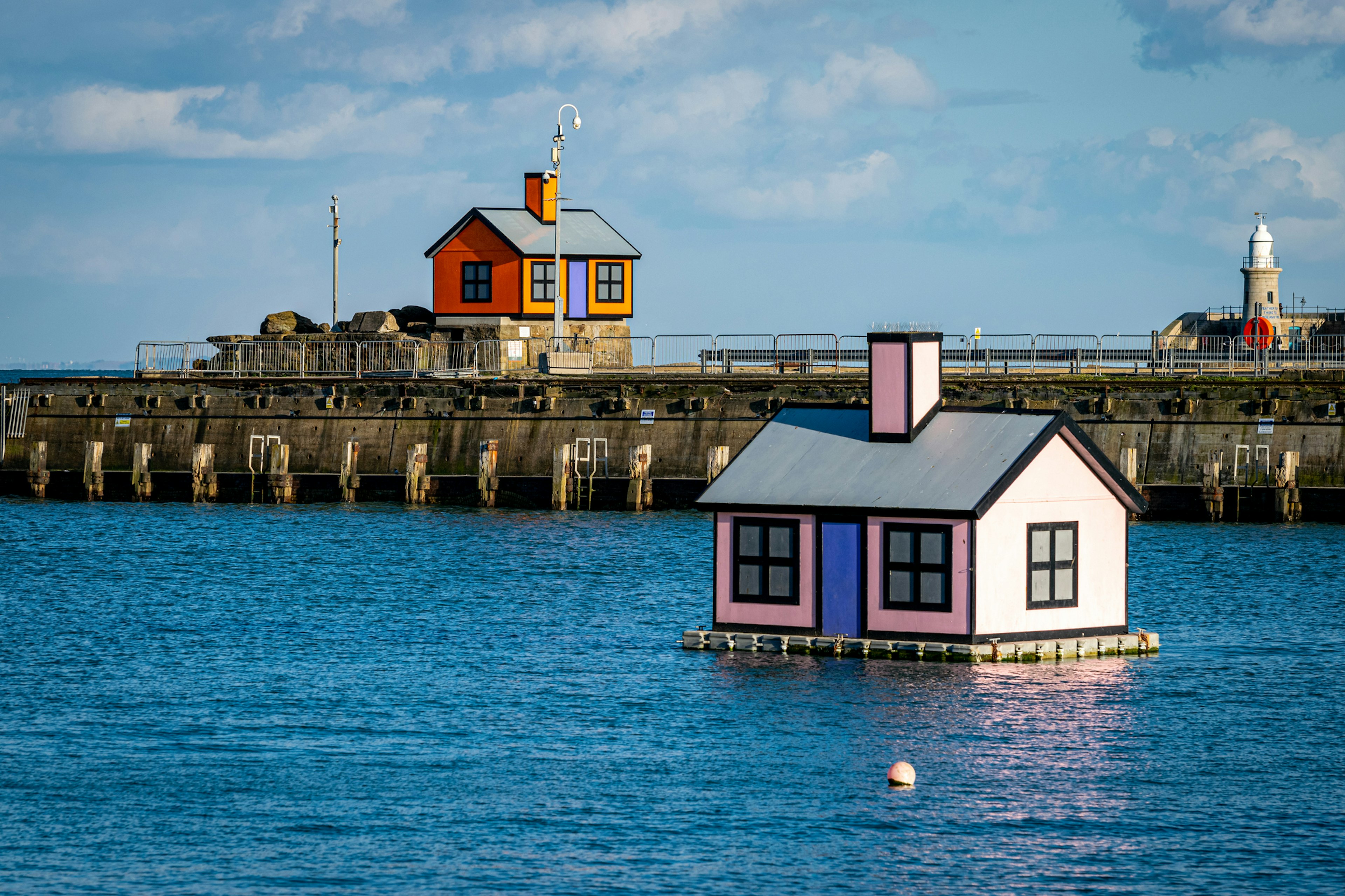 Two instalations of small houses - one floats on a platform on the harbor; the other sits on the harbor arm