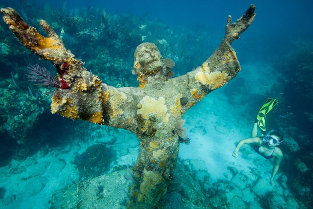 A snorkeler swims by a large underwater statue of Christ with two arms reaching up towards the surface