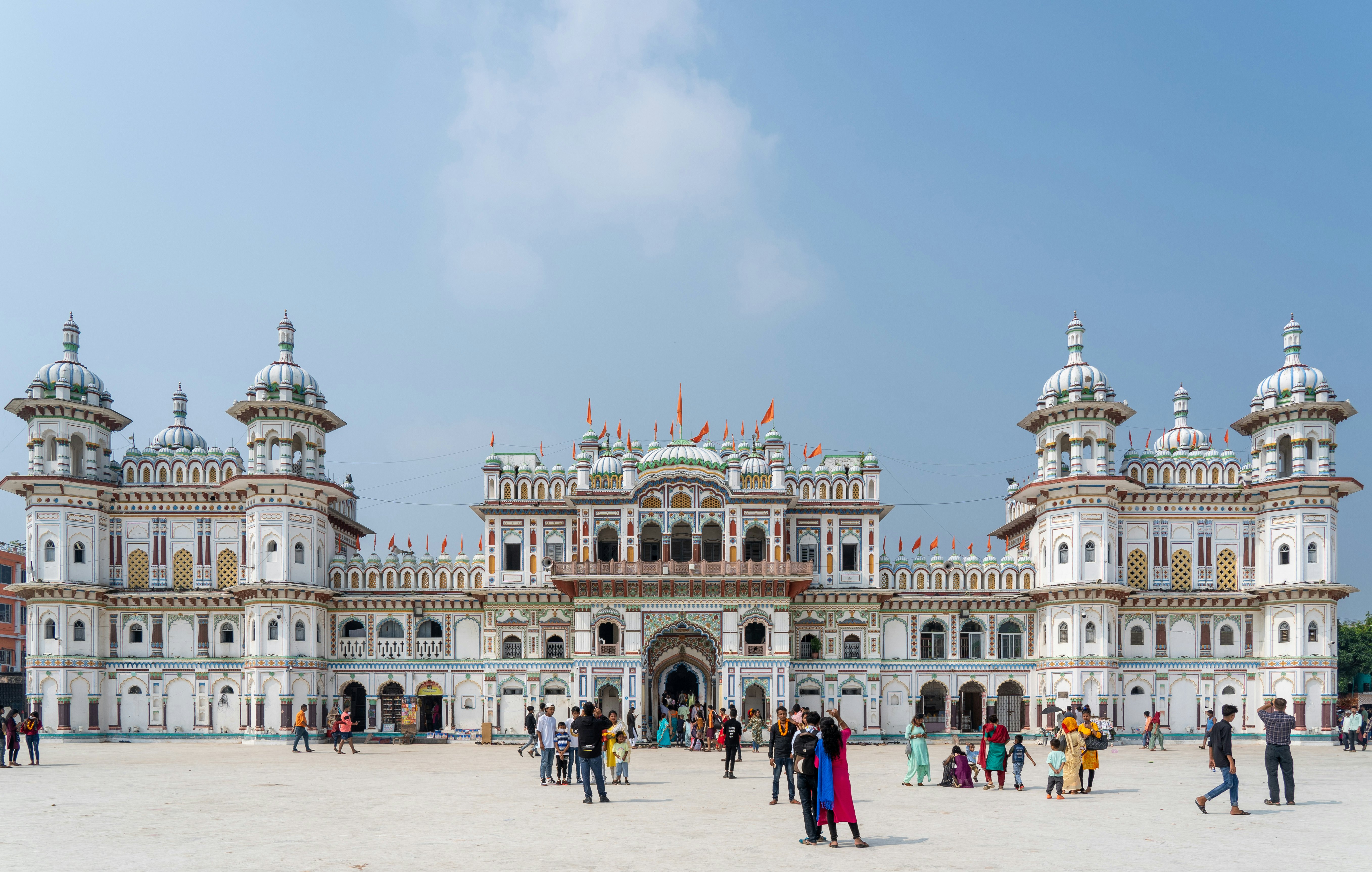 Pilgrims walking on the plaza in front of a vast white temple with domed turrets and orange flags.