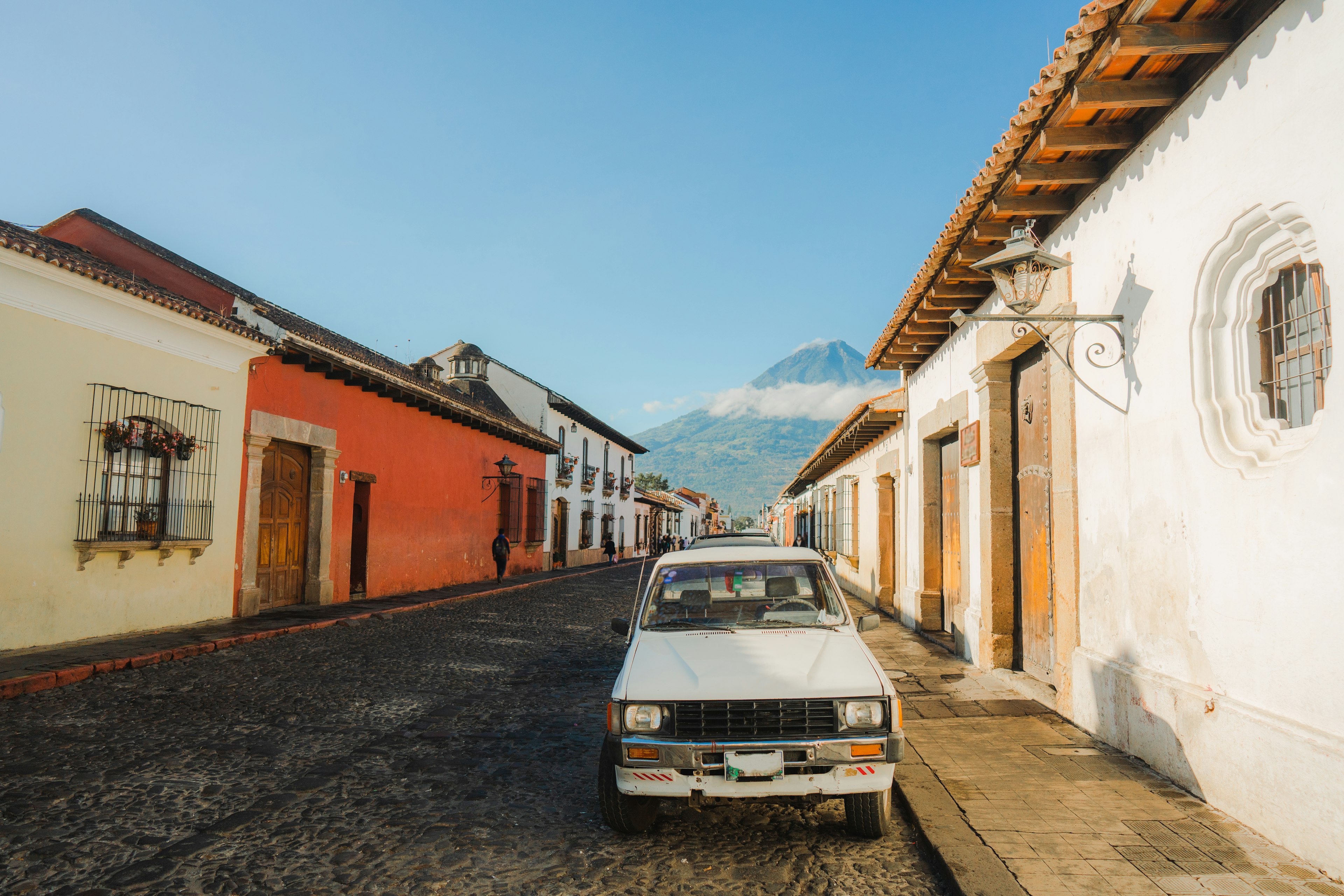 The front of a white pickup truck parked on a cobbled street in Antgua with a mist-shrouded volcano in the distance