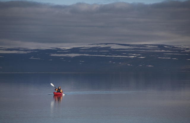 A woman in a red kayak paddles across a very still body of water with snow-capped hills in the distance