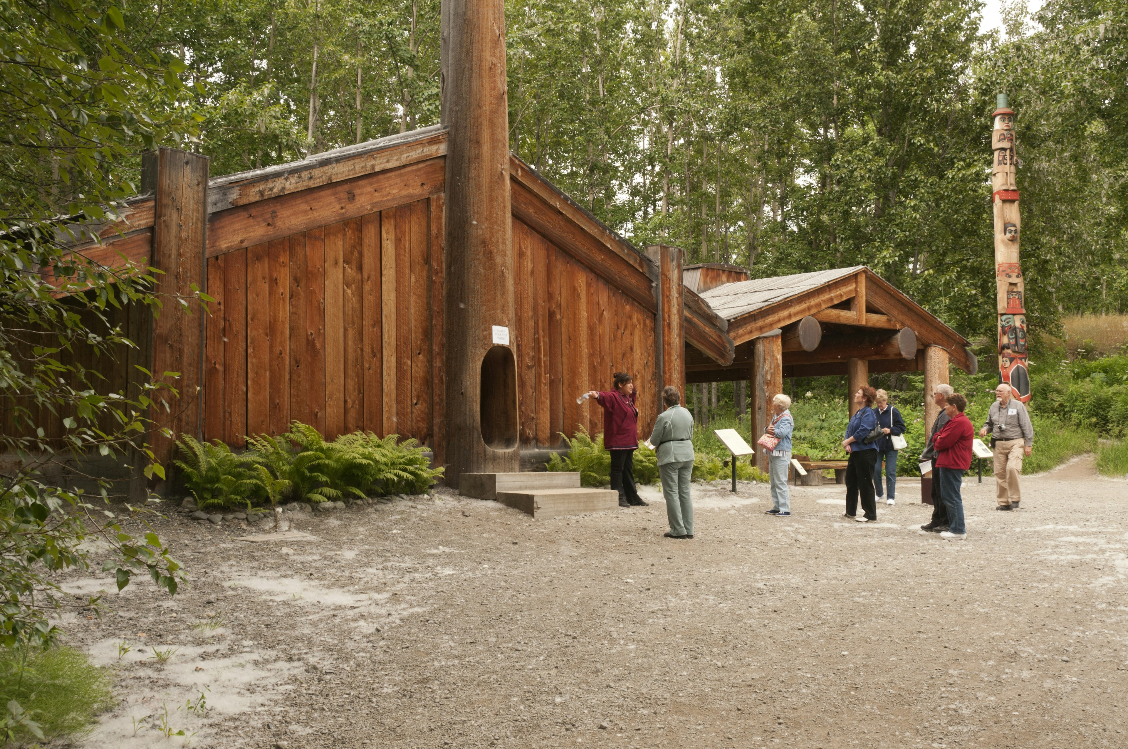 Visitors wander through traditional dwellings in a wooded outdoor area on the grounds of the Alaska Native Heritage Center