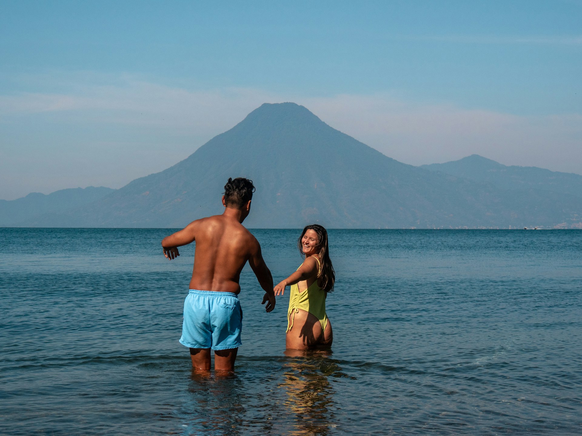 A man and woman in bathing suits wade into the water of Lake Atitlan with a volcano in the distance, Guatemala