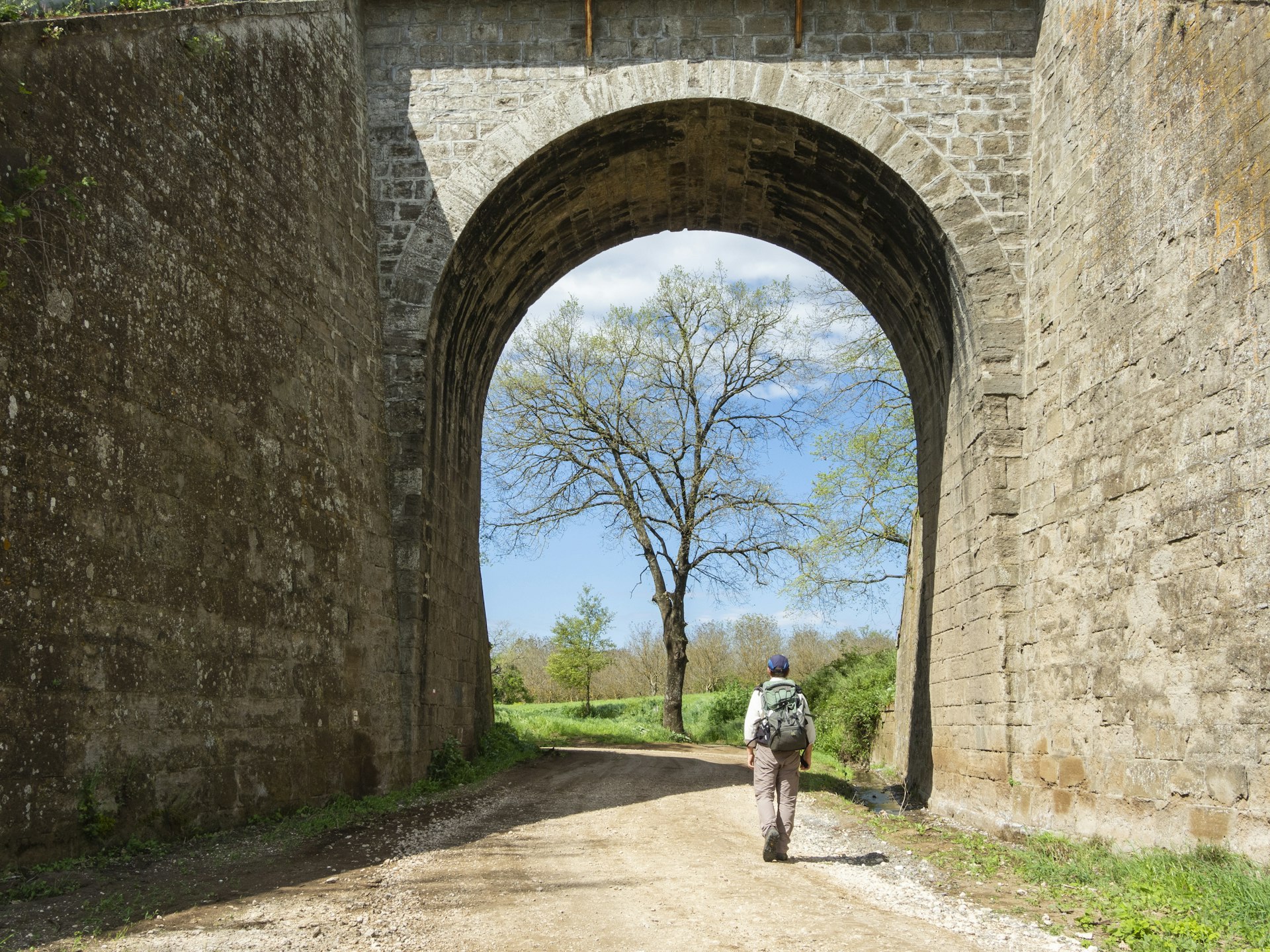 A pilgrim with a backpack walks through a masonry arch along the Via Francigena, near Vetralla