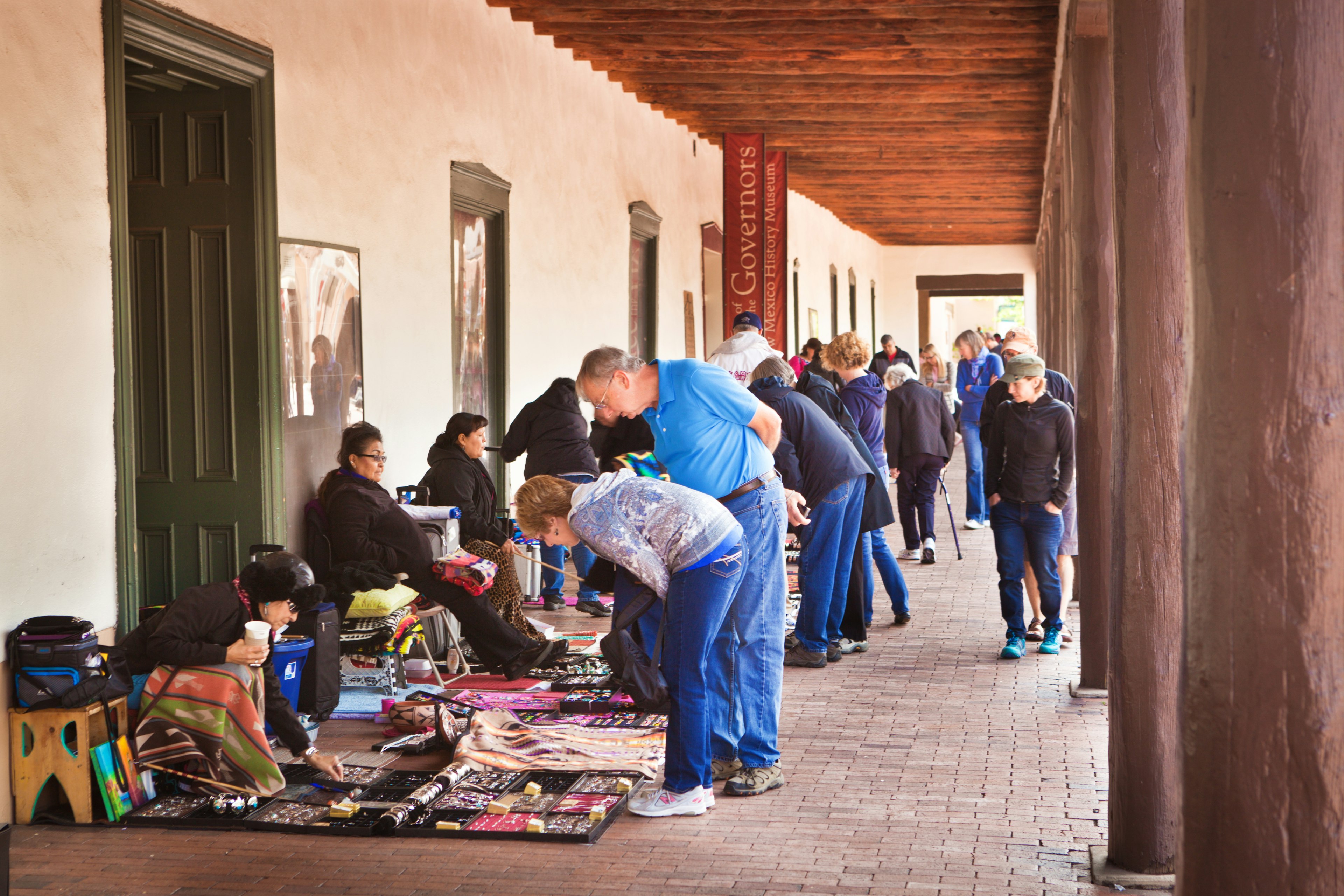 Artists sit near jewlery and other handmade creations for sale in an arcade while tourists look on at what they might buy