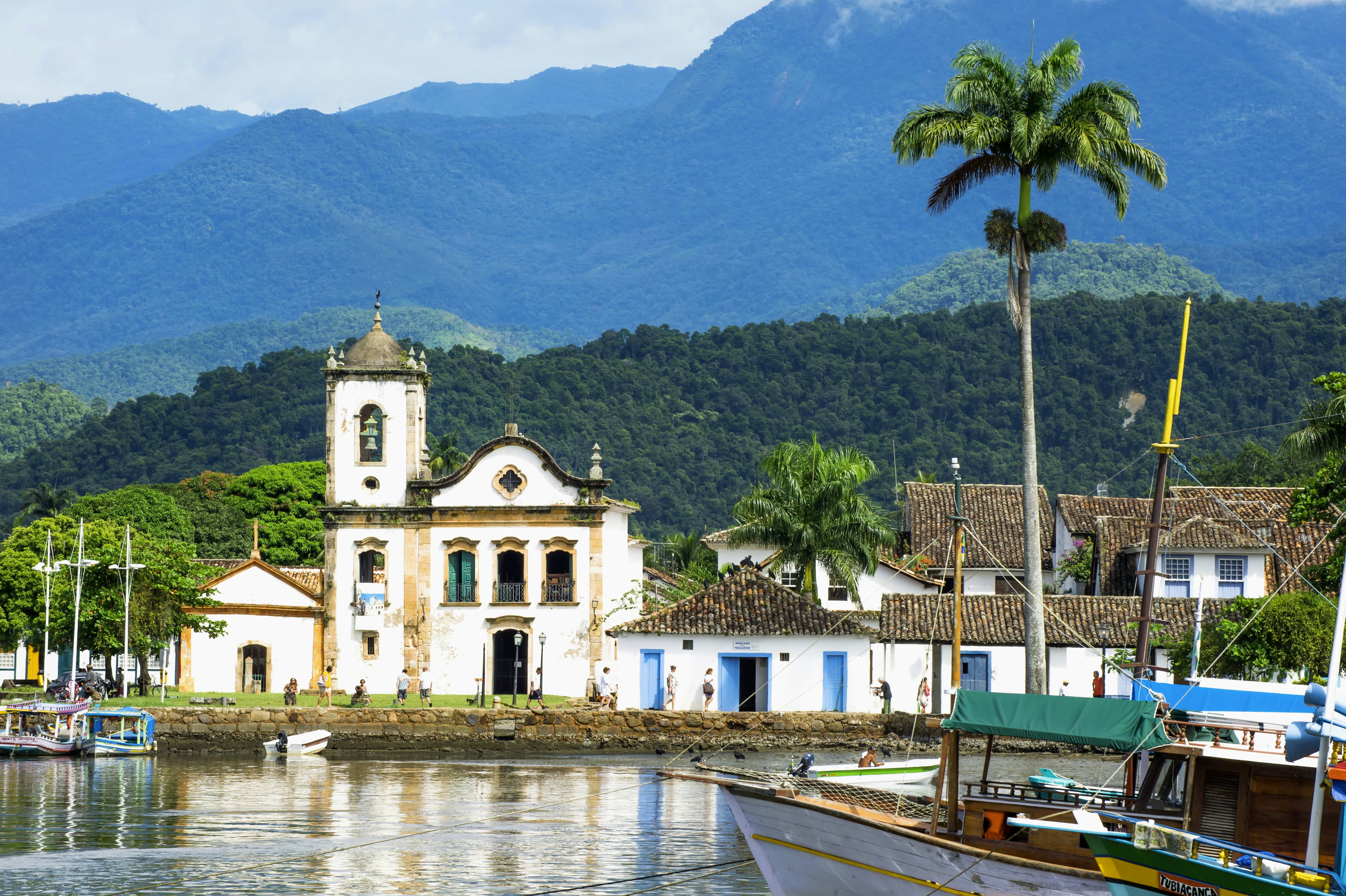 A Portuguese-style white church with a bell tower faces the water, with palm trees and green mountains beyond.