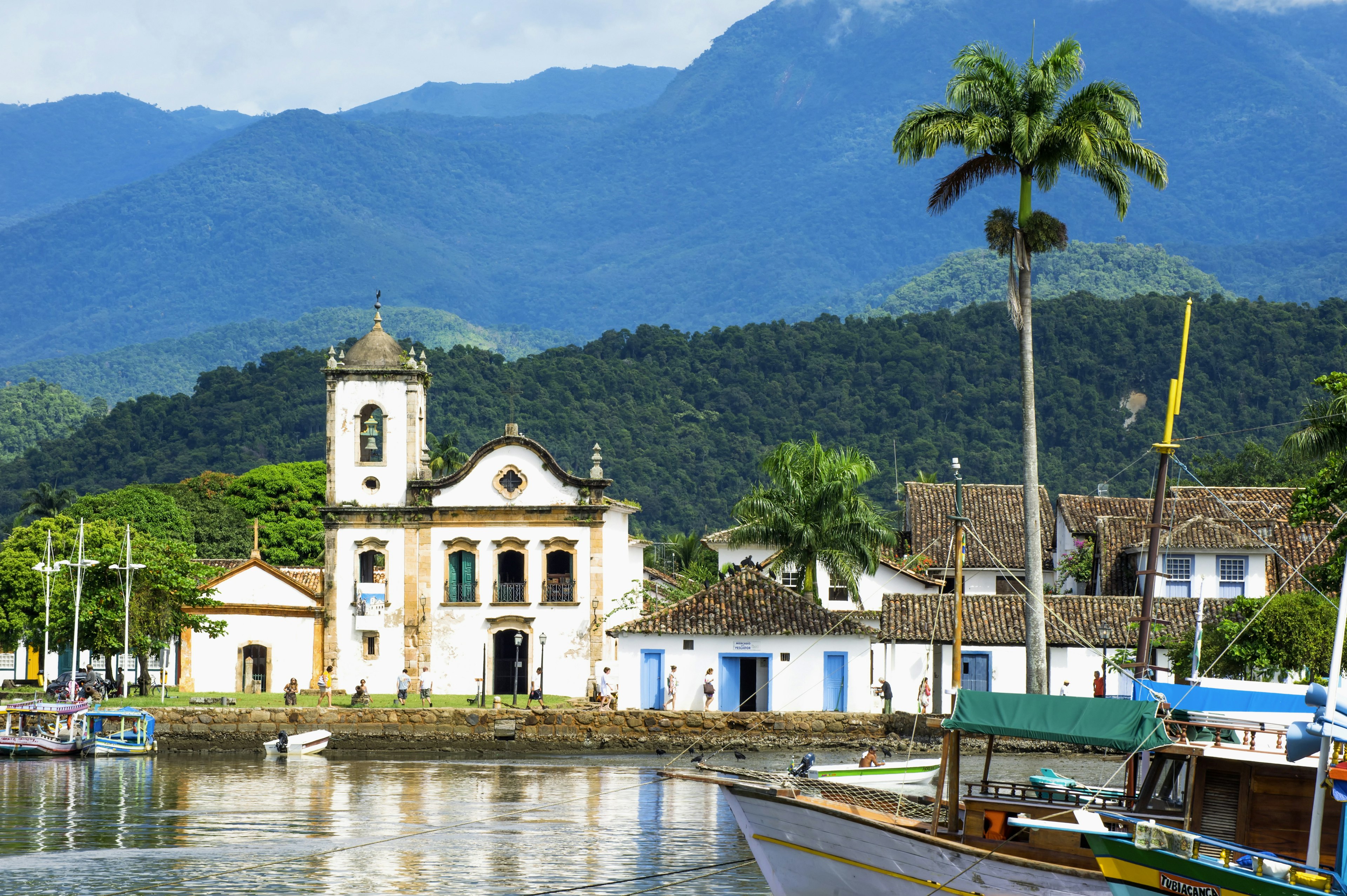 A colonial-era church in Paraty, Brazil, seen from the water, with palm trees and green mountains beyond