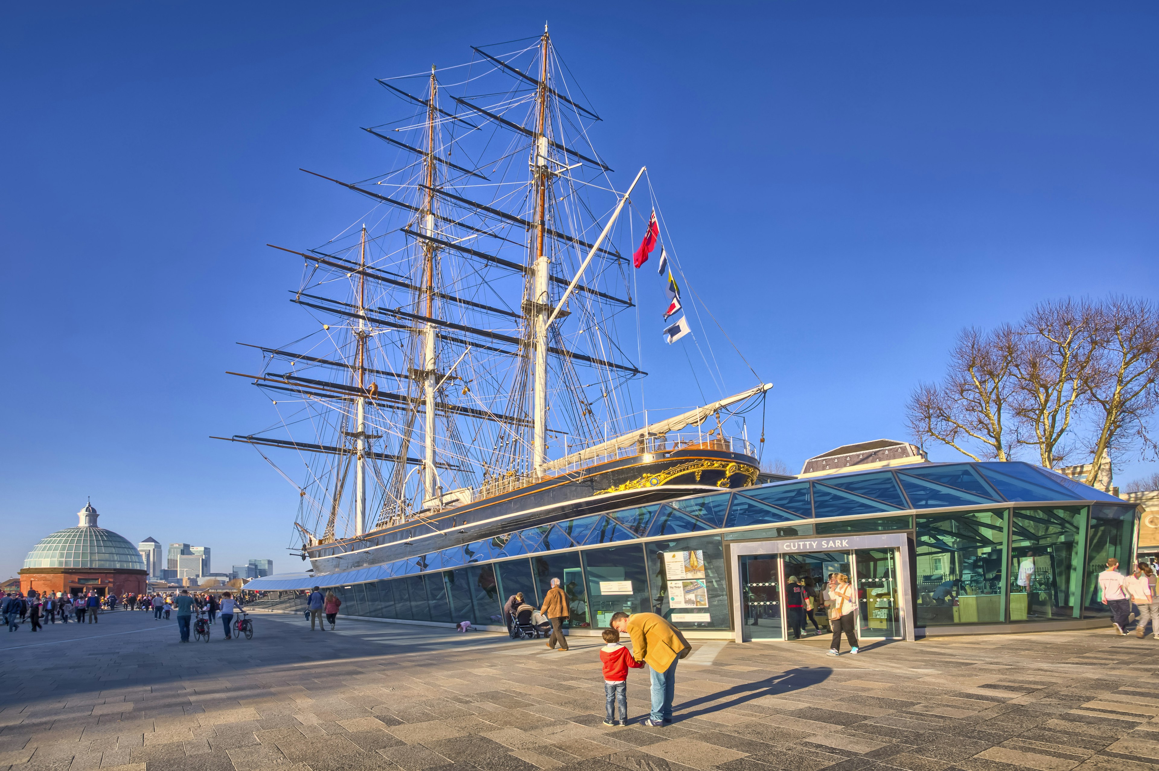 A tea clipper boat on land surrounded by glass to form a museum building. A man kneels down next to a child in the foreground, pointing upwards at the boat's masts.