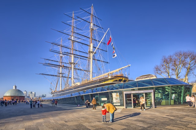 A tea clipper boat on land surrounded by glass to form a museum building. A man kneels down next to a child in the foreground, pointing upwards at the boat's masts.