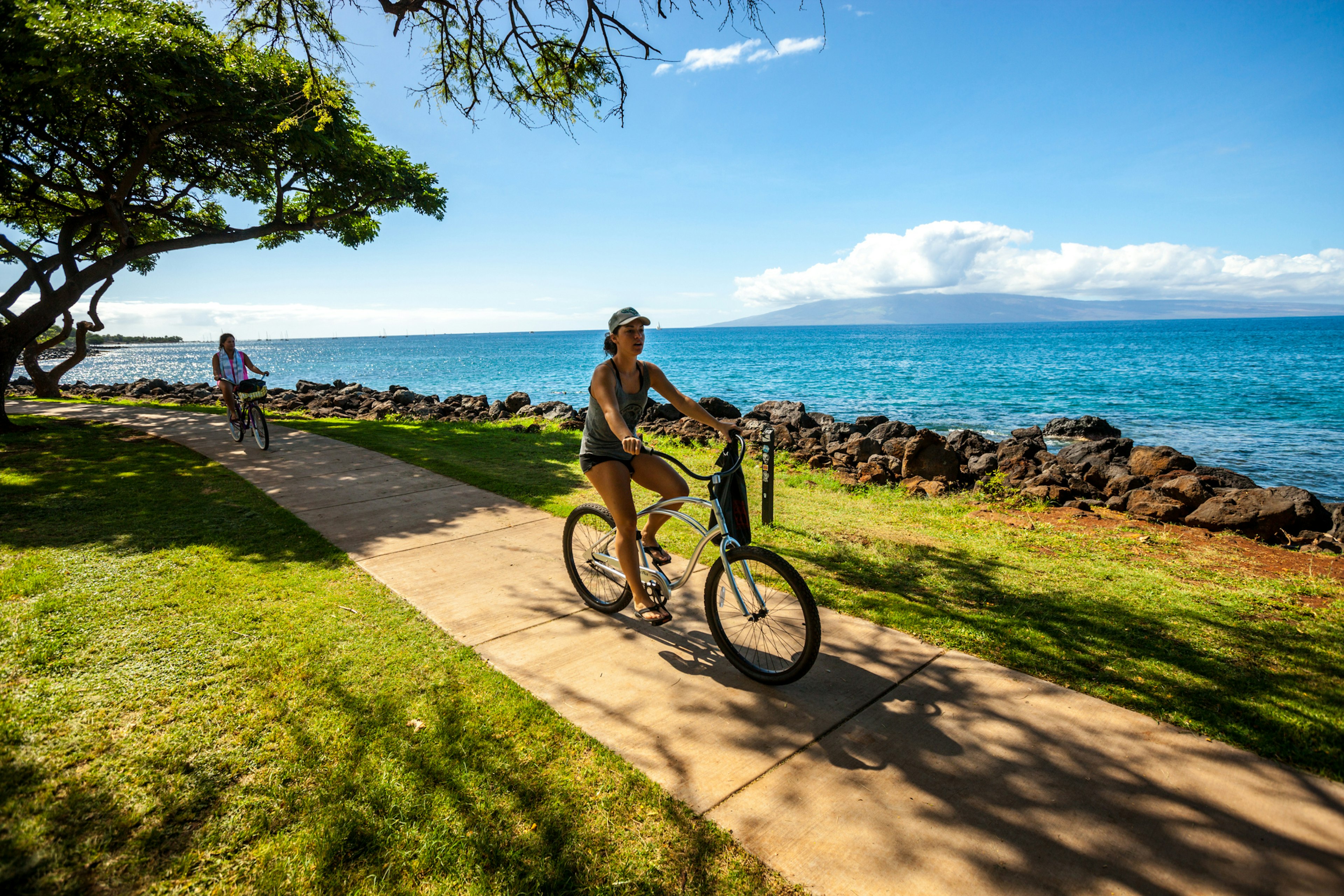 Two women ride bicycles along a paved path that runs parallel to the sea