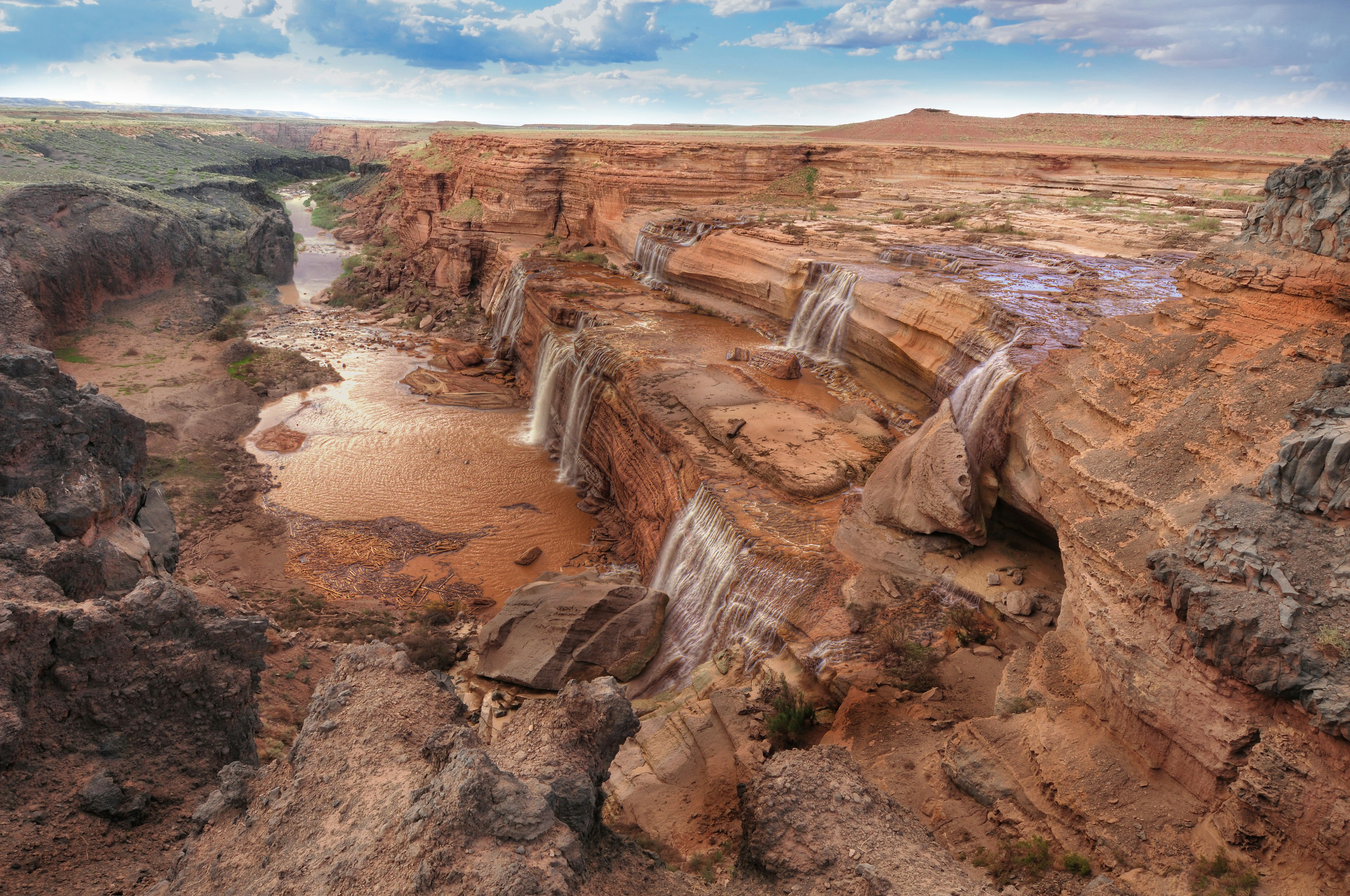 Several small cascades flow down to a river off a red-brown rocky formation