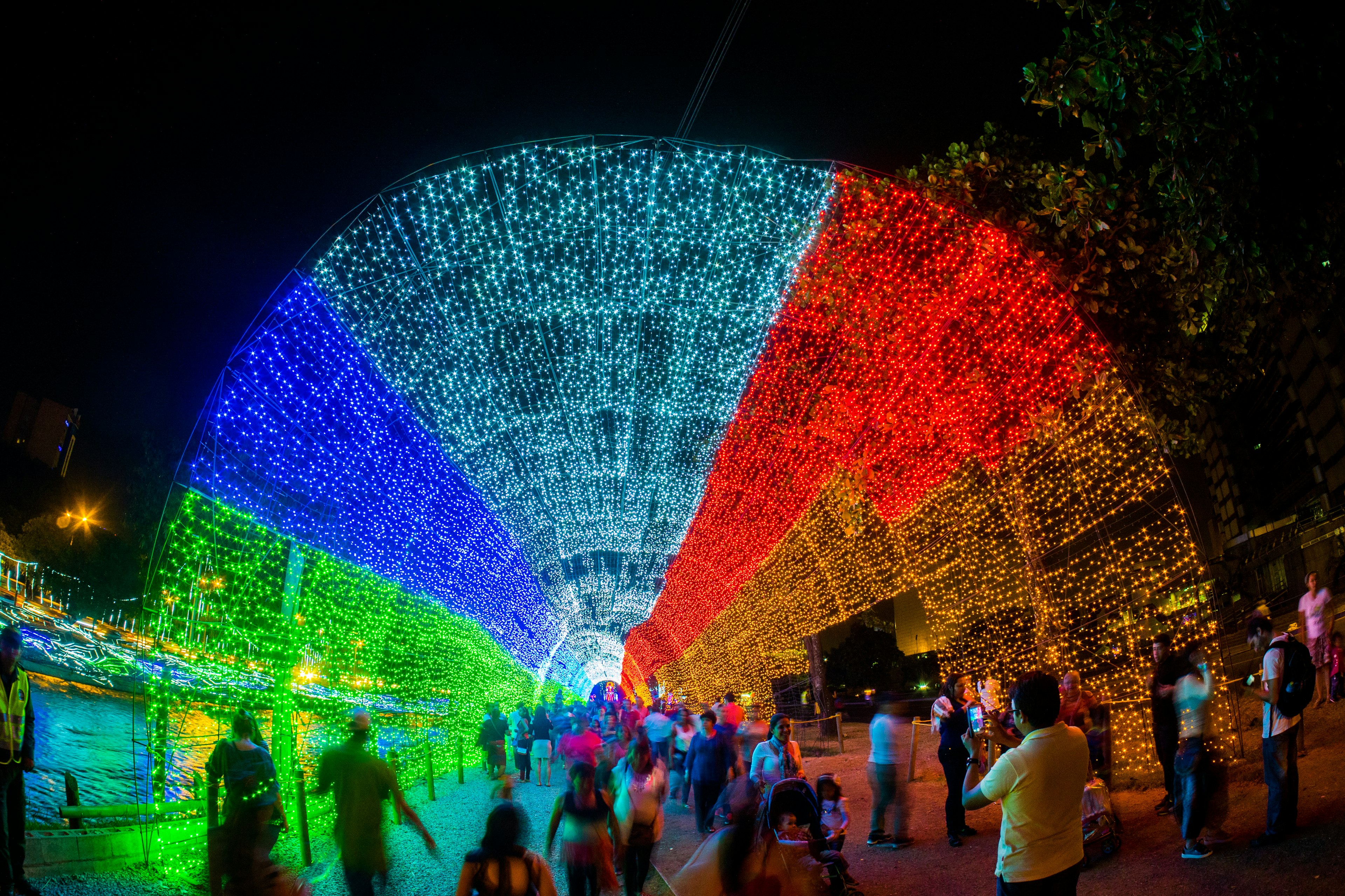 A tunnel of brightly colored lights loops over a pedestrian street at night. People gather to admire the lights and take photos