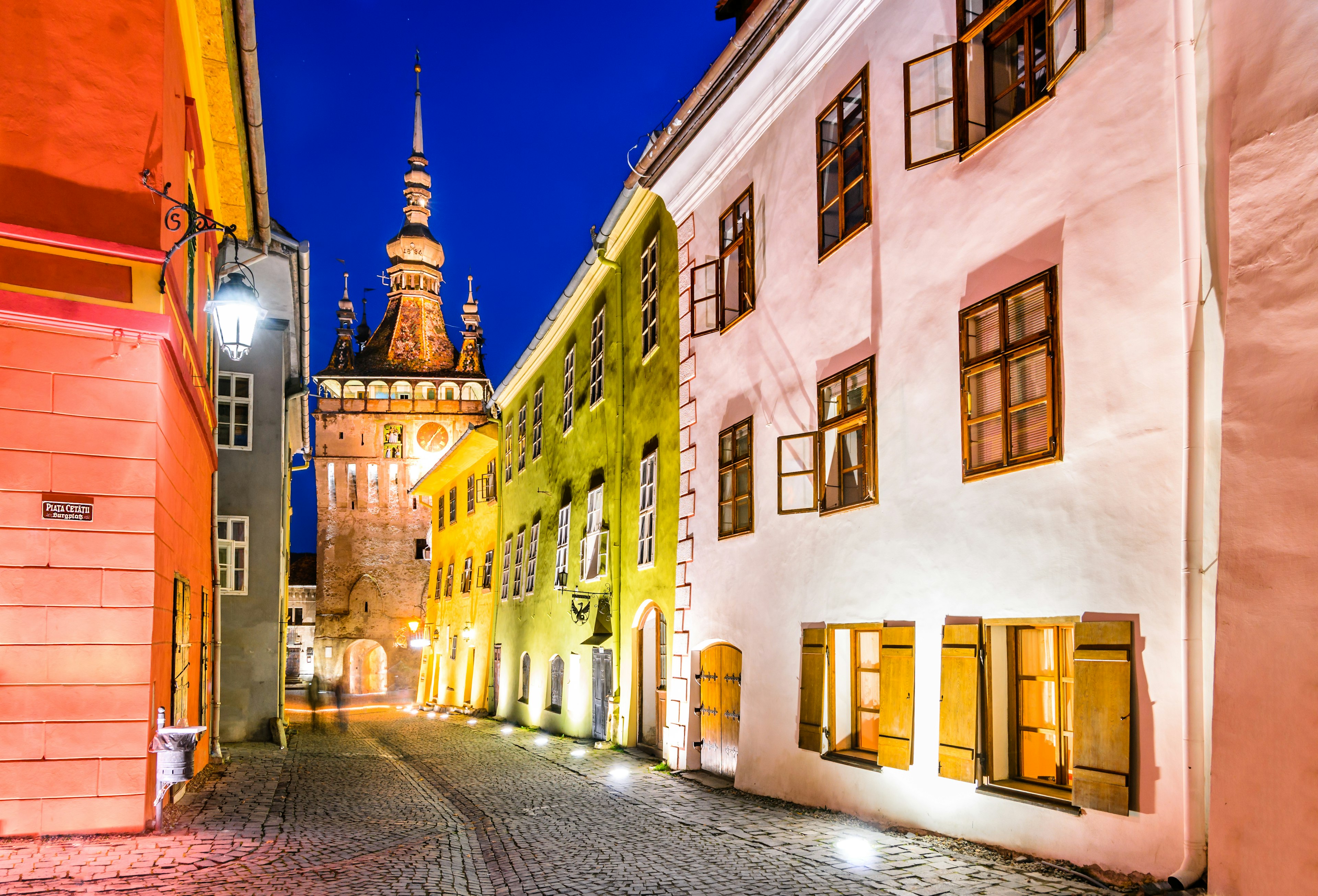 A view of a cobbled street with historic buildings illuminated by night, with a clock tower in the distance