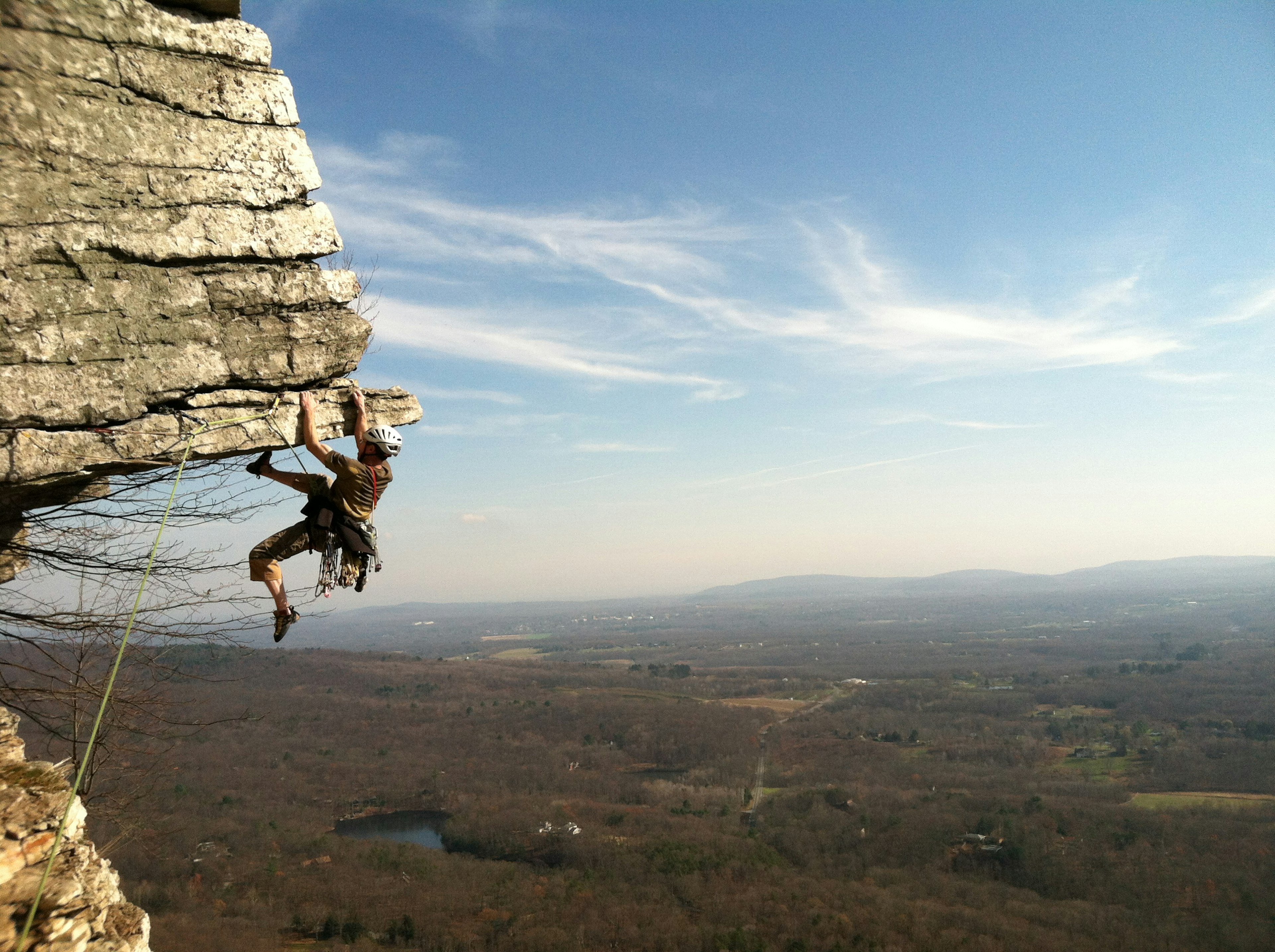 A rock climber tackles a challenging overhang with dense woodland spread out in the distance