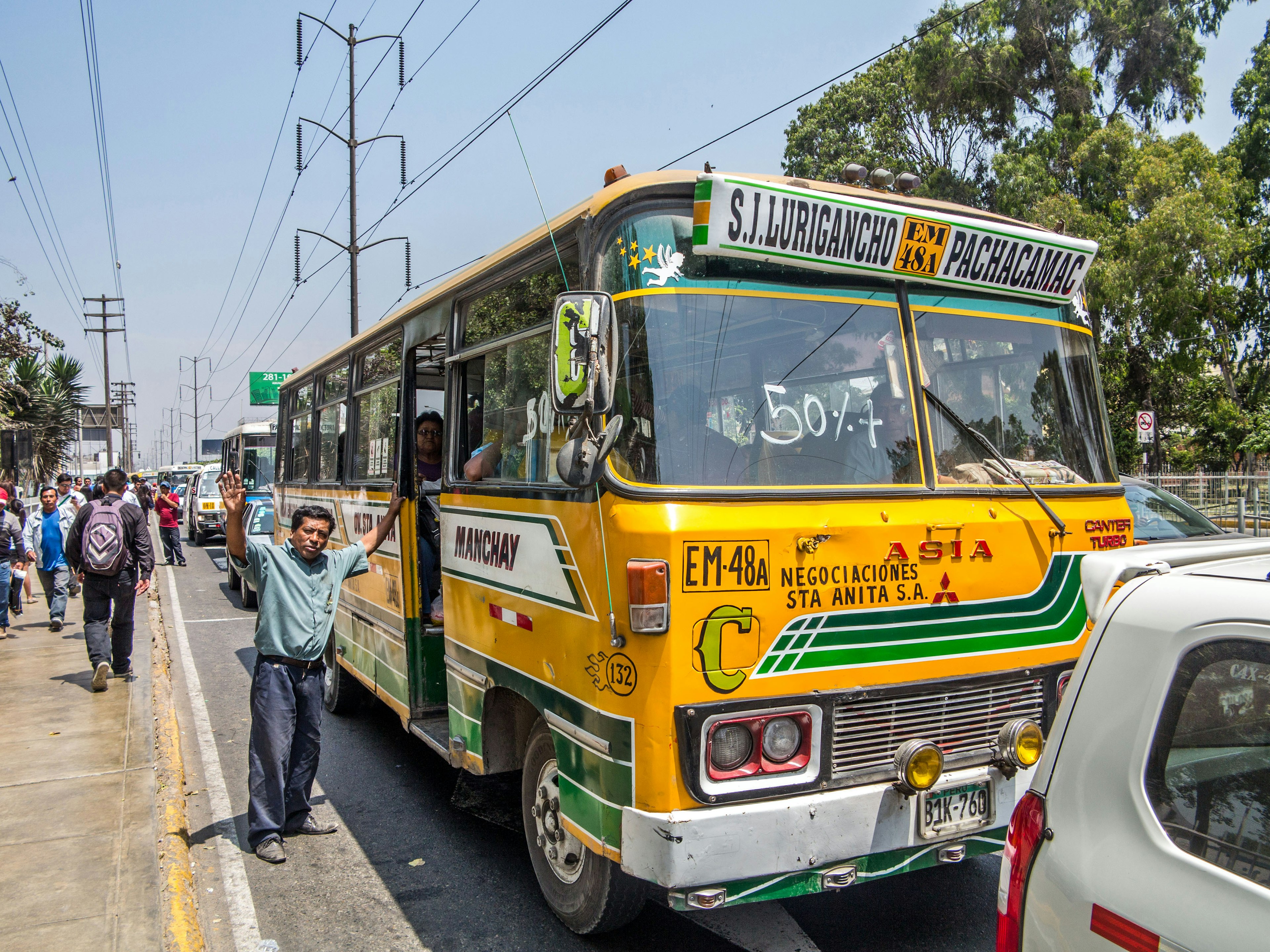 Typical green and yellow public transport bus in Lima street. Driver is visible behind the windscreen and the ticket collector is standing outside of the entrance door. Pedestrians pictured in the background