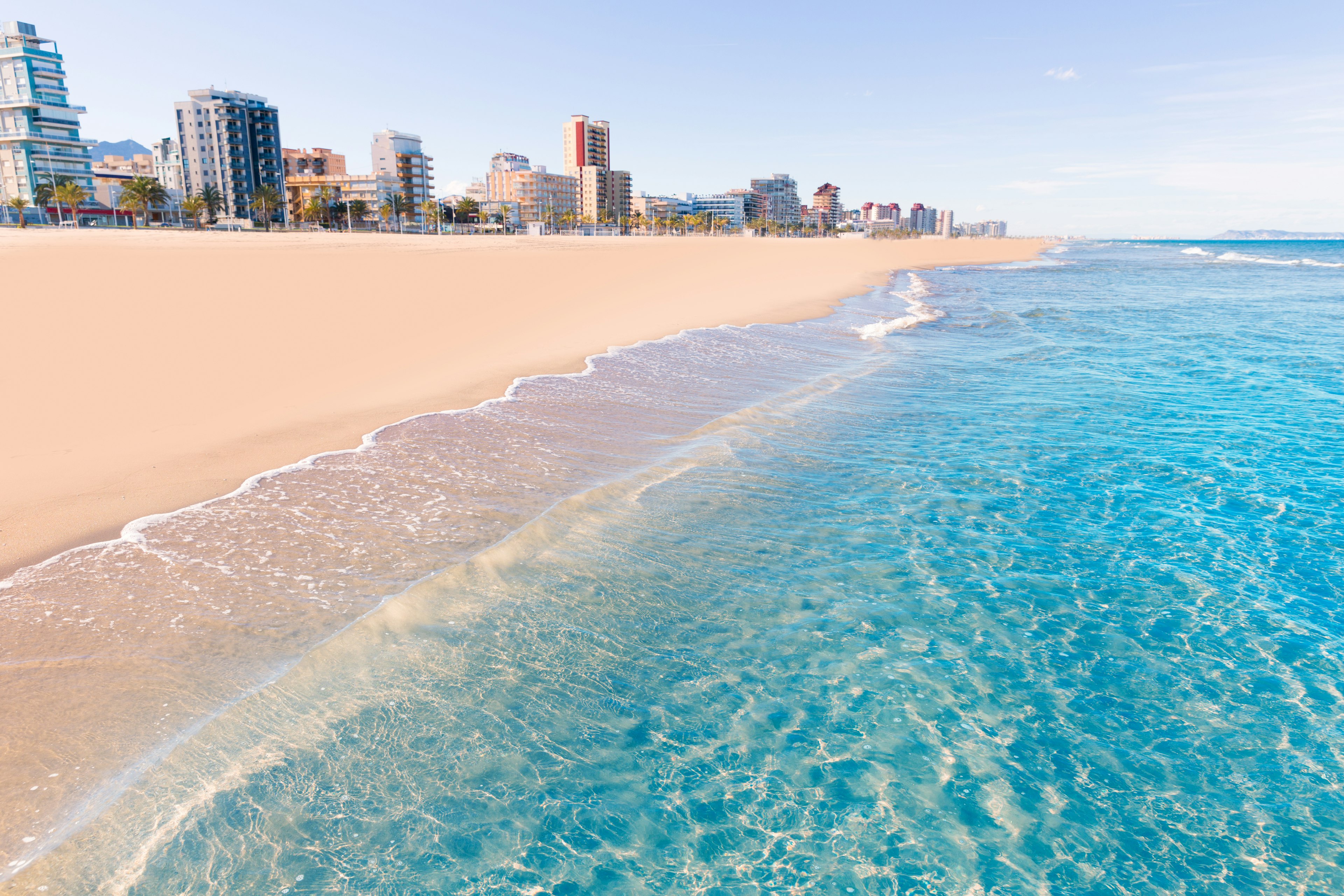 The clear, blue wash of the Mediterranean Sea cuts diagonally against the soft, pinkish sands of Playa de Gandia in Valencia, Spain with several tower blocks rising up in the background