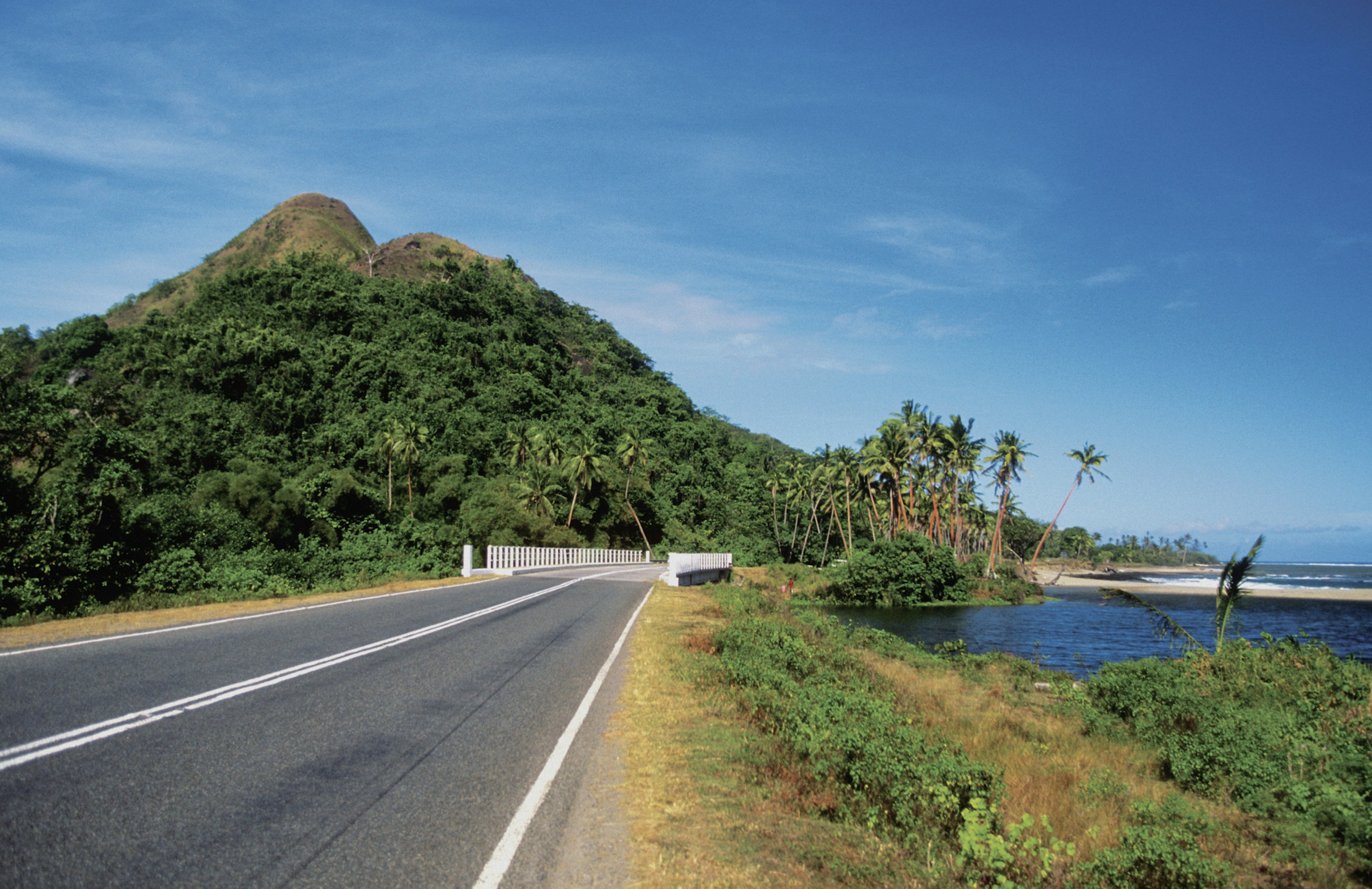 An empty coastal highway with a sandy palm-lined beach to right and a hill in the distance on Viti Levu, Fiji