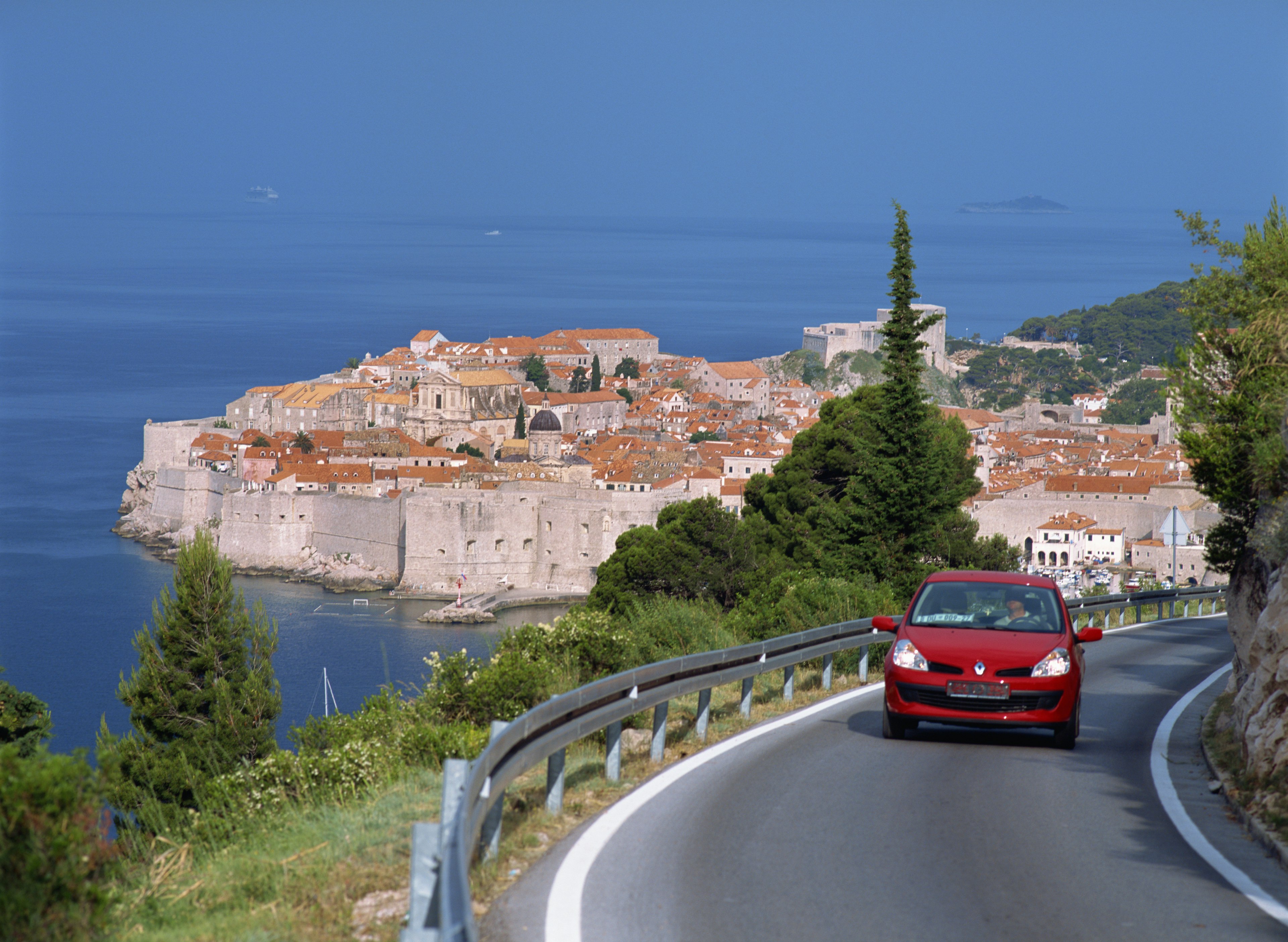 A red car driving around a bend with Dubrovnik in the background