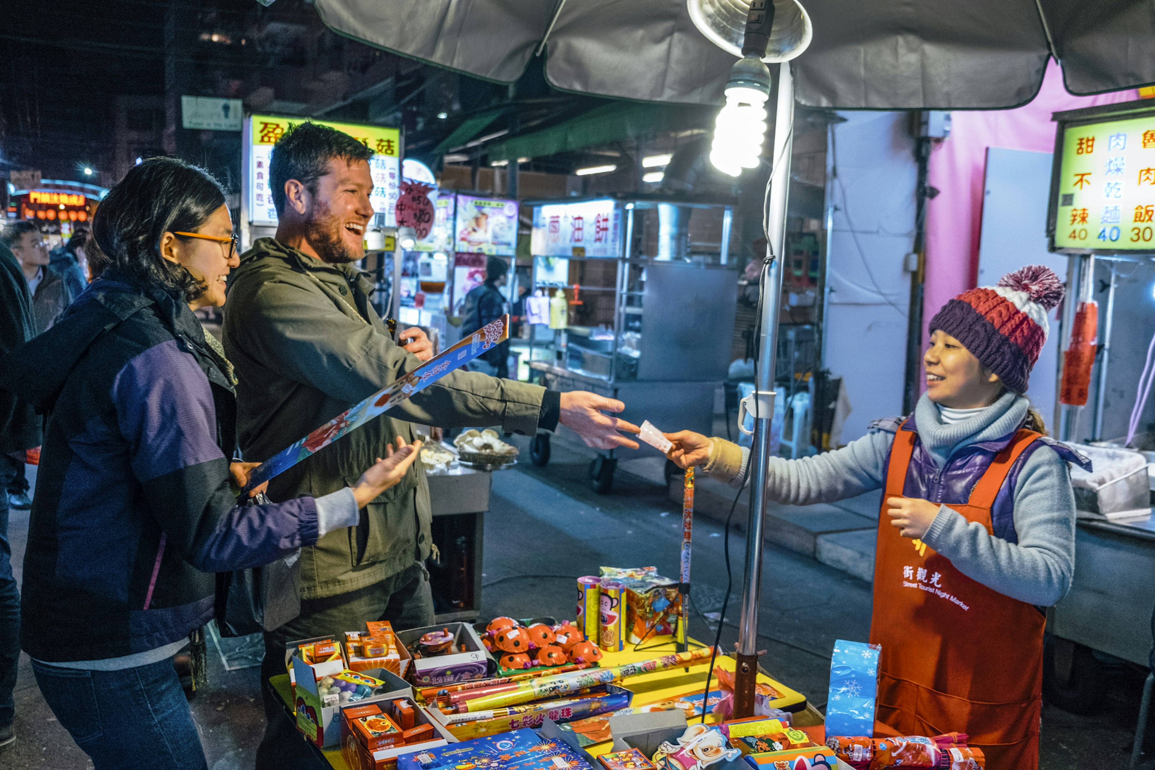 Two people smile as they hand over money to a vendor at a night market stall