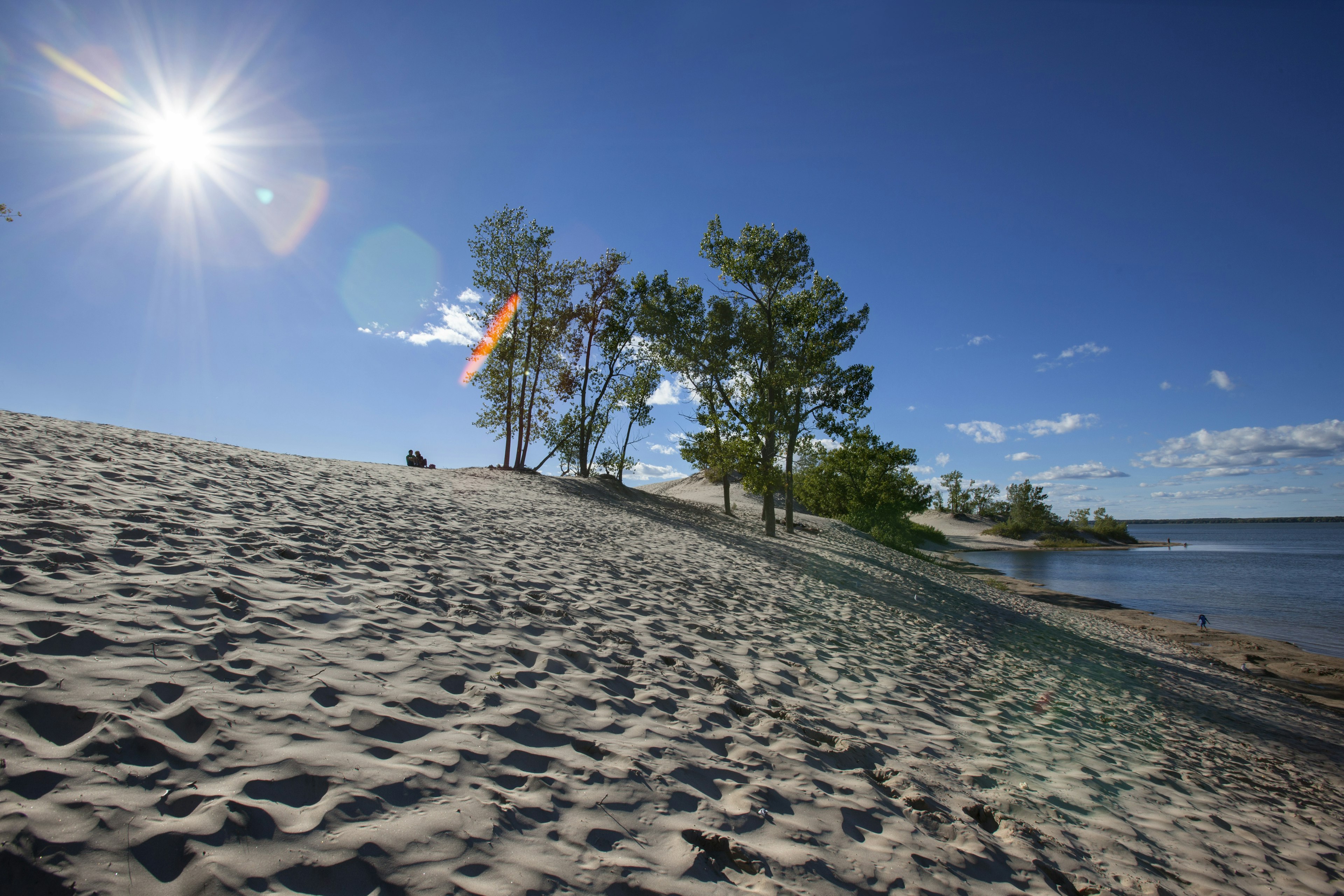 Two people sit at the top of a sand dune looking down towards a lake