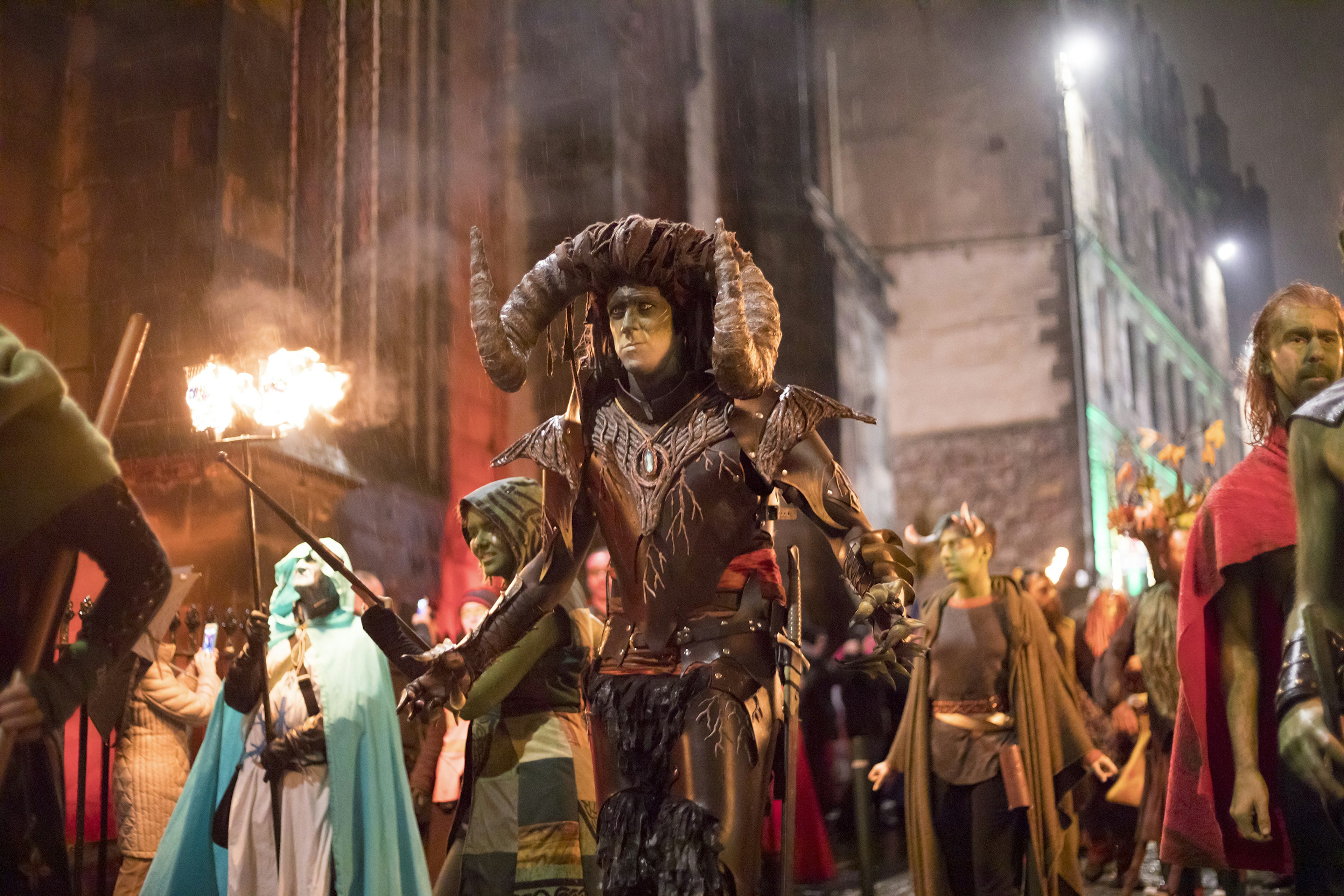 People in intricate costume walk the streets of a city. Some are holding burning lanterns high above them